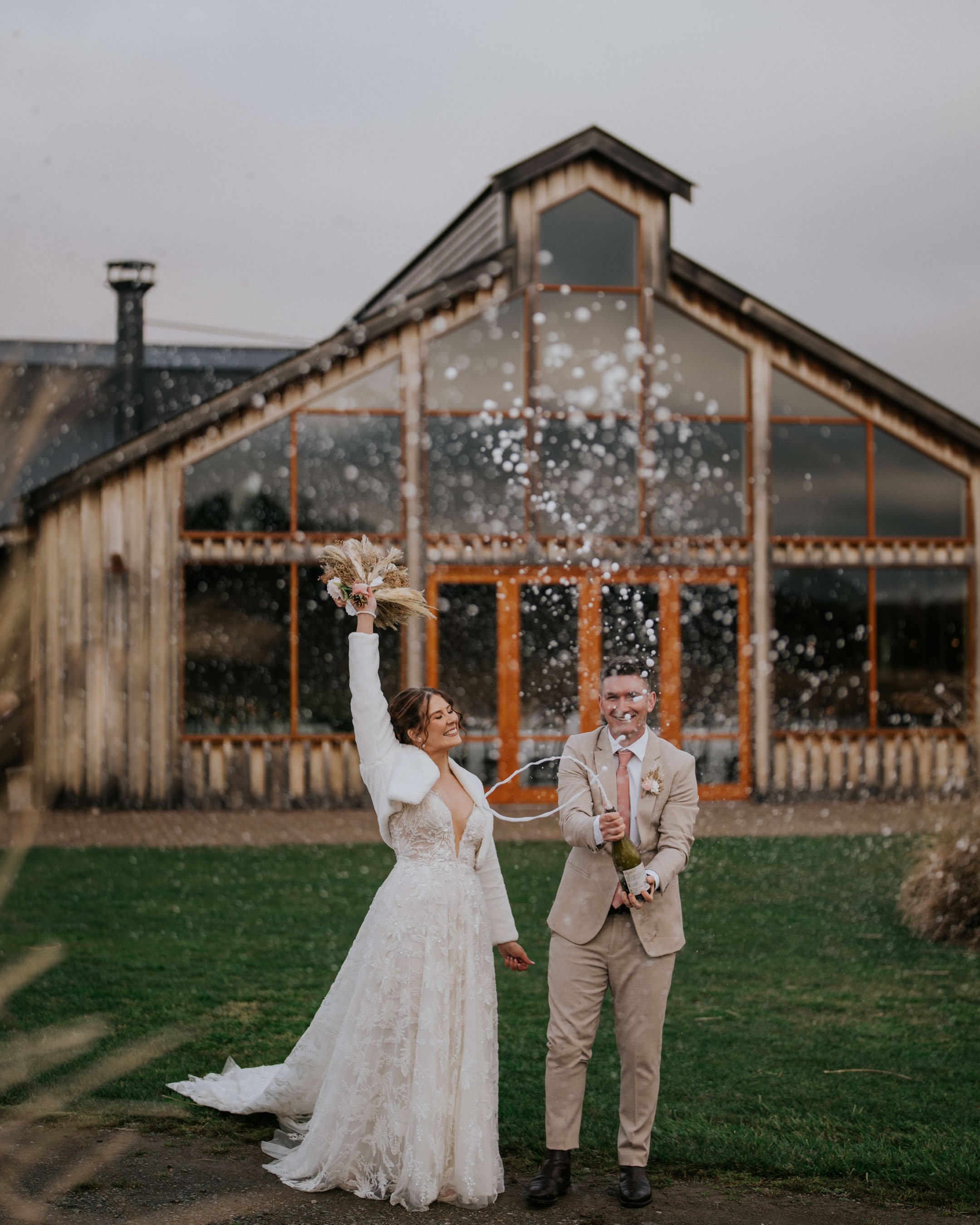 A bride and groom celebrating outdoors in front of a barn-like building, with the bride holding a bouquet and the groom opening a bottle of champagne, spraying it in celebration.