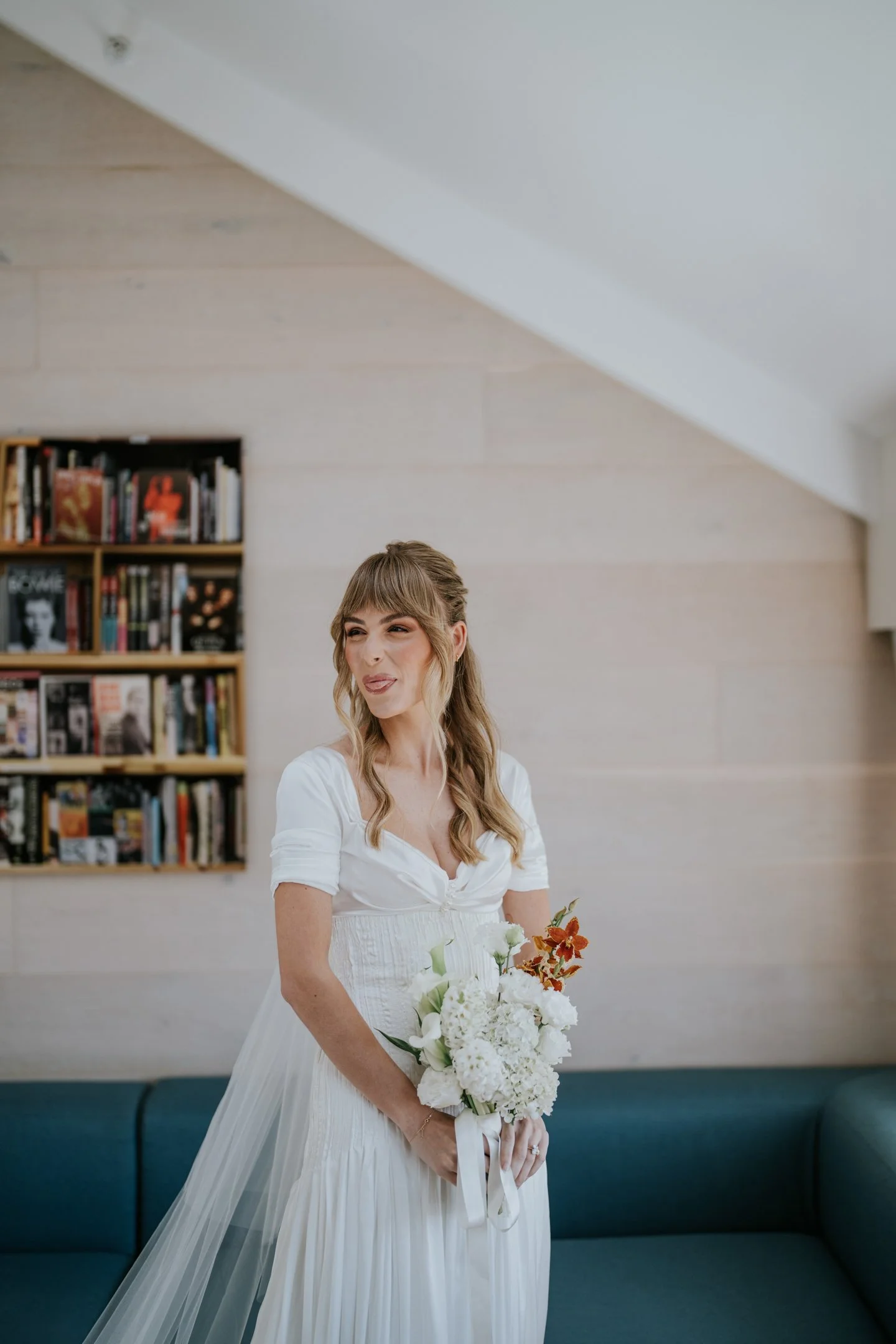 A woman in a white dress holding a bouquet of flowers, standing in a room with a bookshelf filled with books and a blue cushioned bench against the wall.