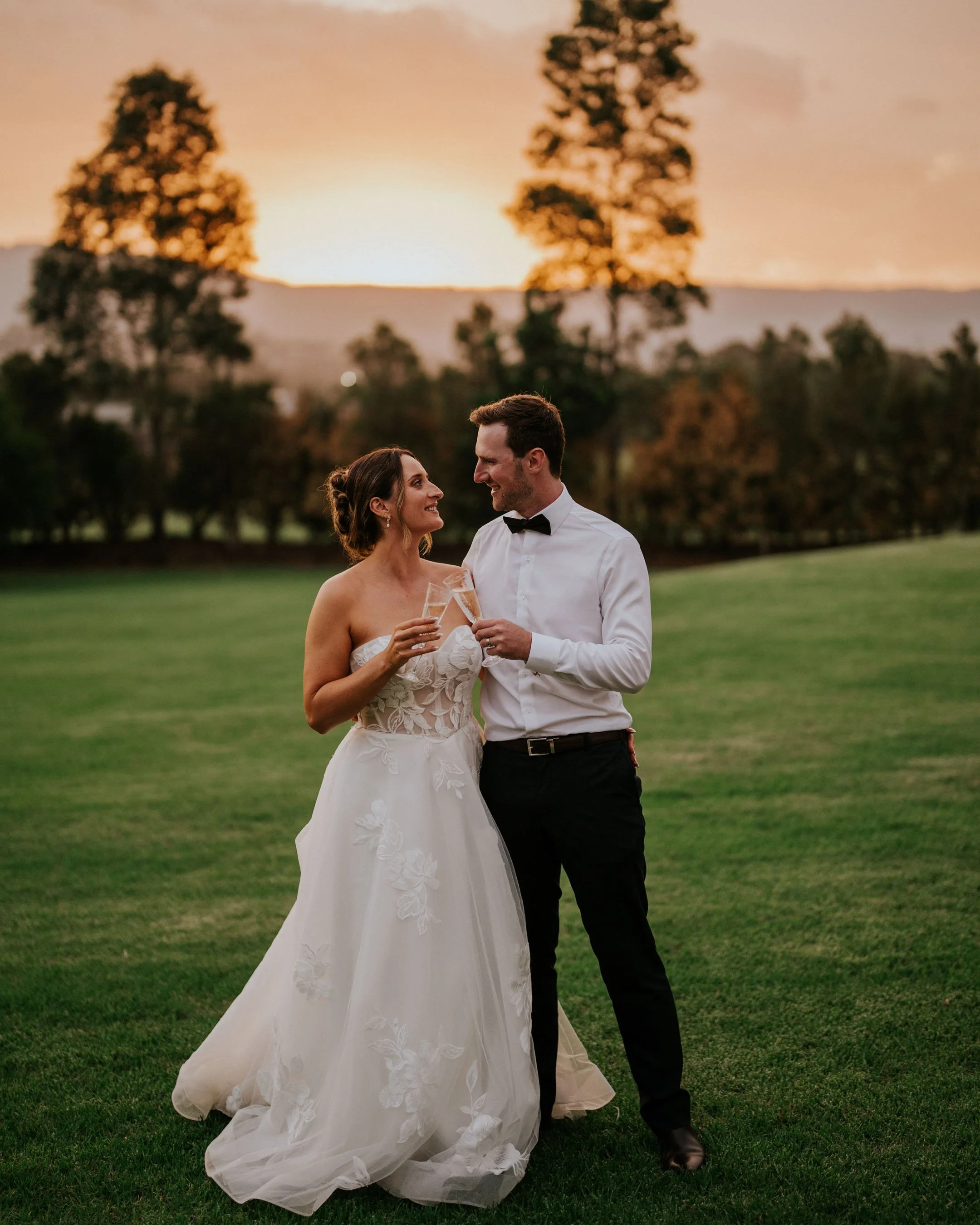 A bride and groom standing on a grassy field during sunset, smiling and holding glasses, with trees and hills in the background.