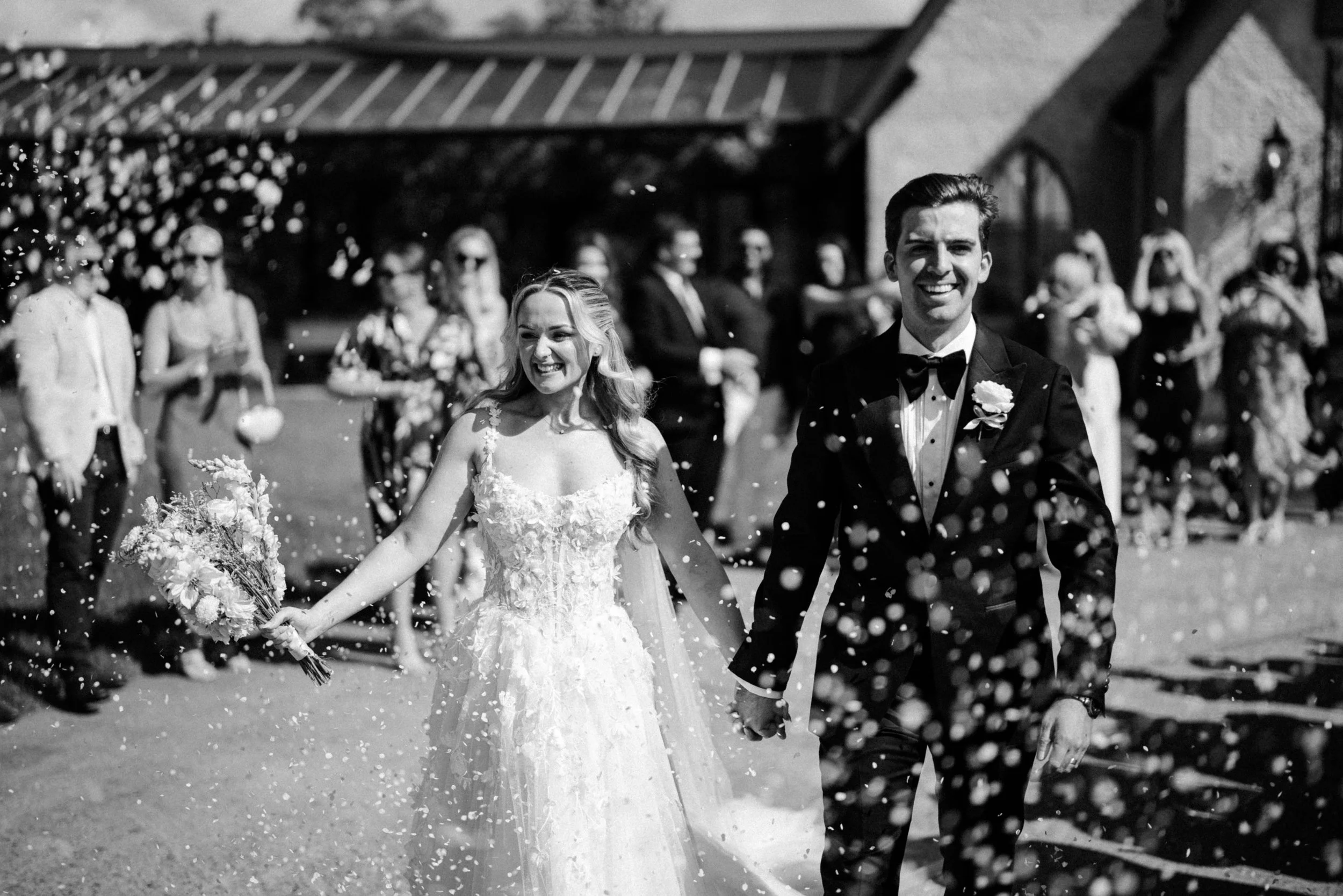 A black-and-white photograph of a newlywed couple holding hands and smiling as they walk through a shower of confetti, surrounded by friends and family at an outdoor wedding ceremony.