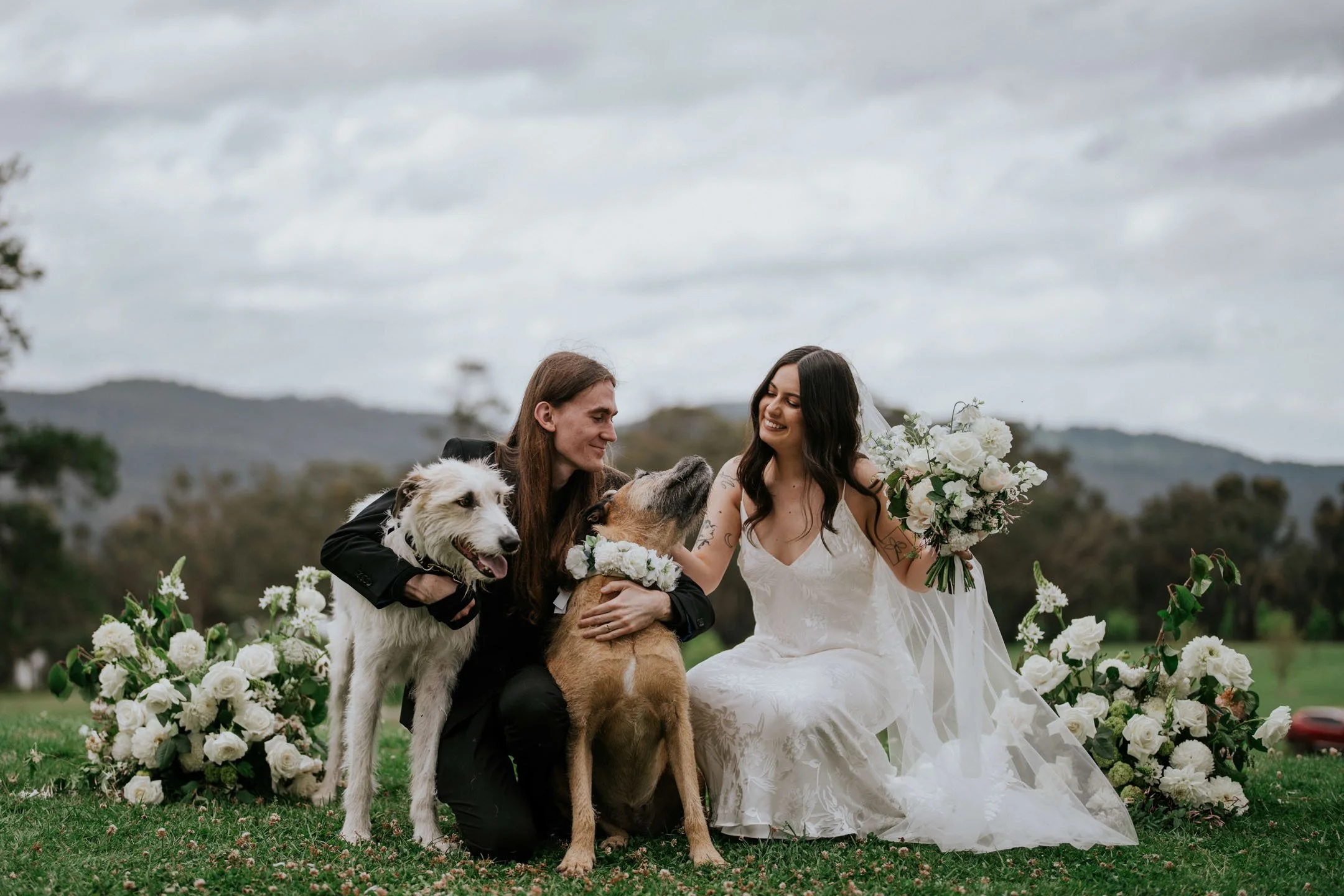 A wedding scene outdoors showing a bride and a groom with three dogs, surrounded by white floral arrangements, with a cloudy sky and distant hills in the background.