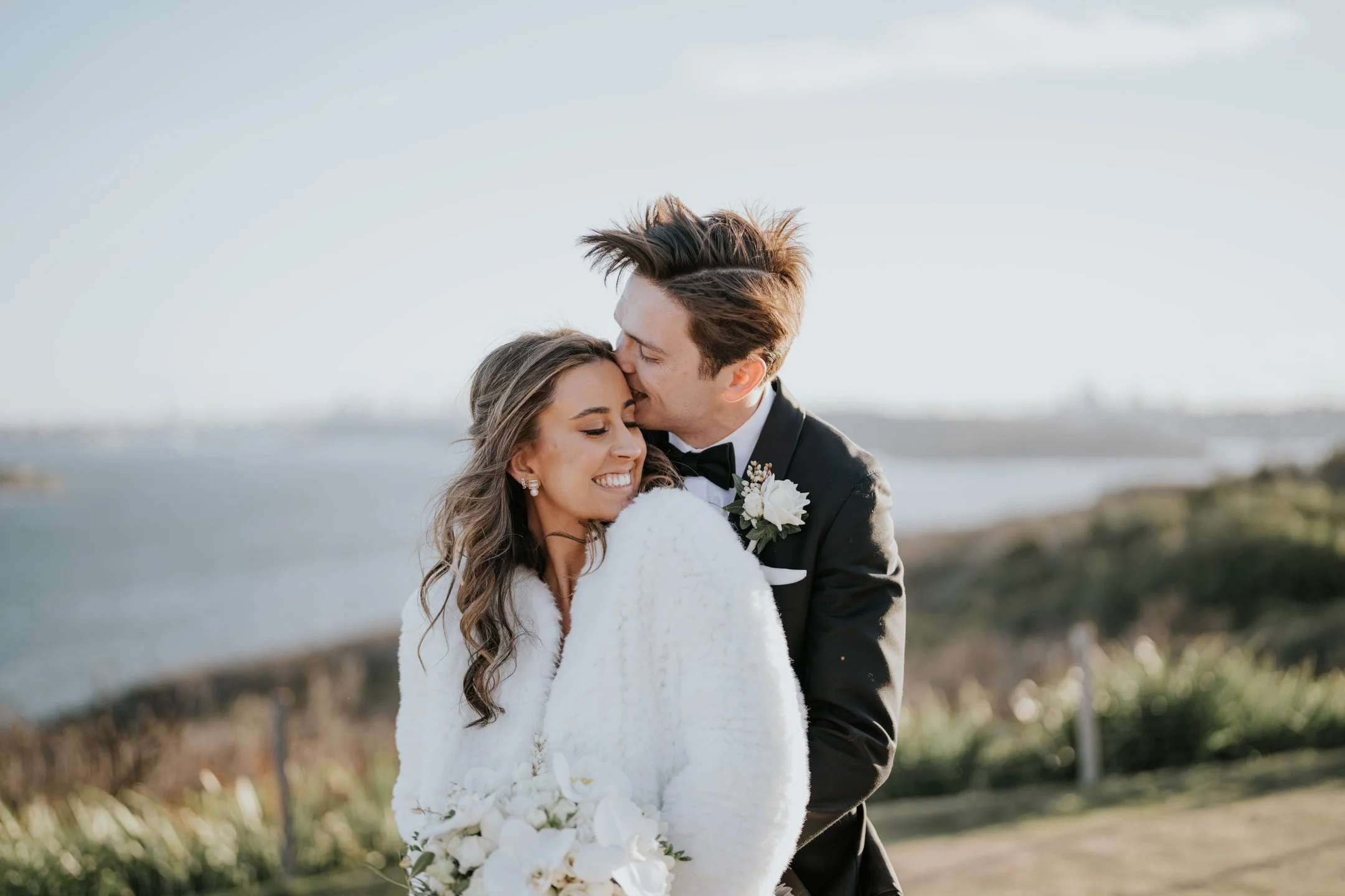 A happy couple on their wedding day, with the groom kissing the bride's forehead and holding her close in an outdoor setting near a body of water.