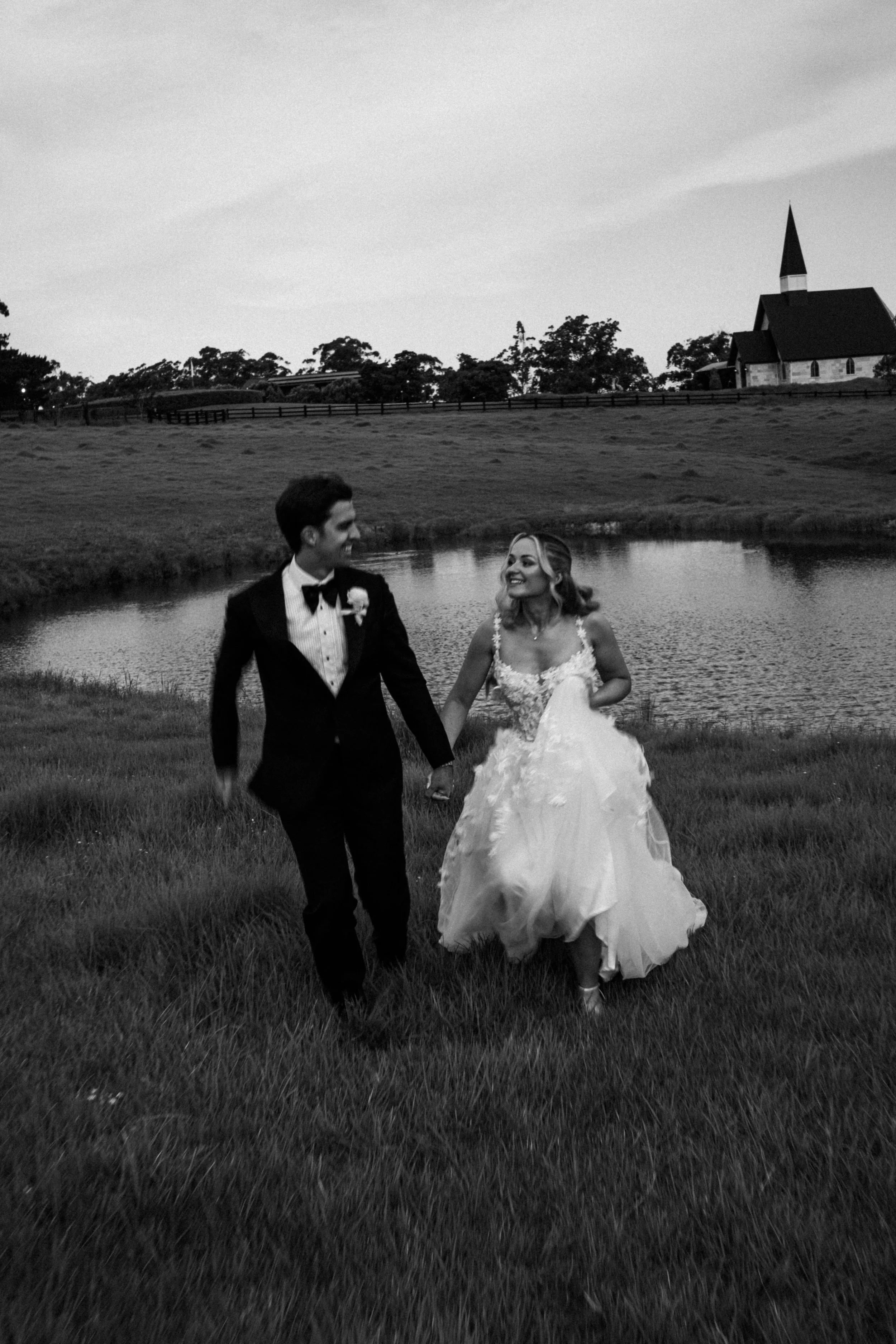 A black and white photo of a bride and groom holding hands and smiling at each other near a pond with a church and trees in the background.