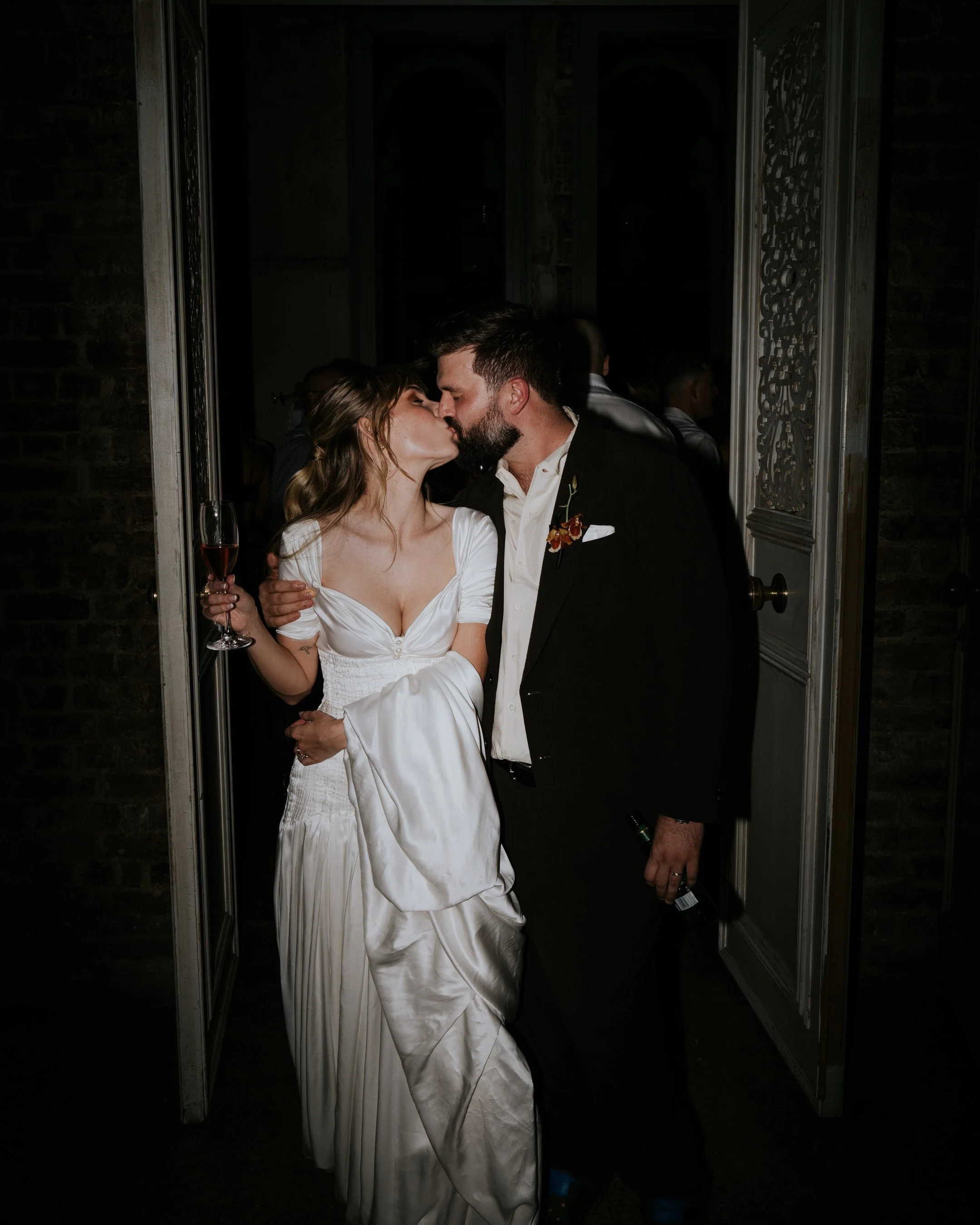 A couple in wedding attire sharing a kiss at their wedding reception, framed by an open door with people in the background.