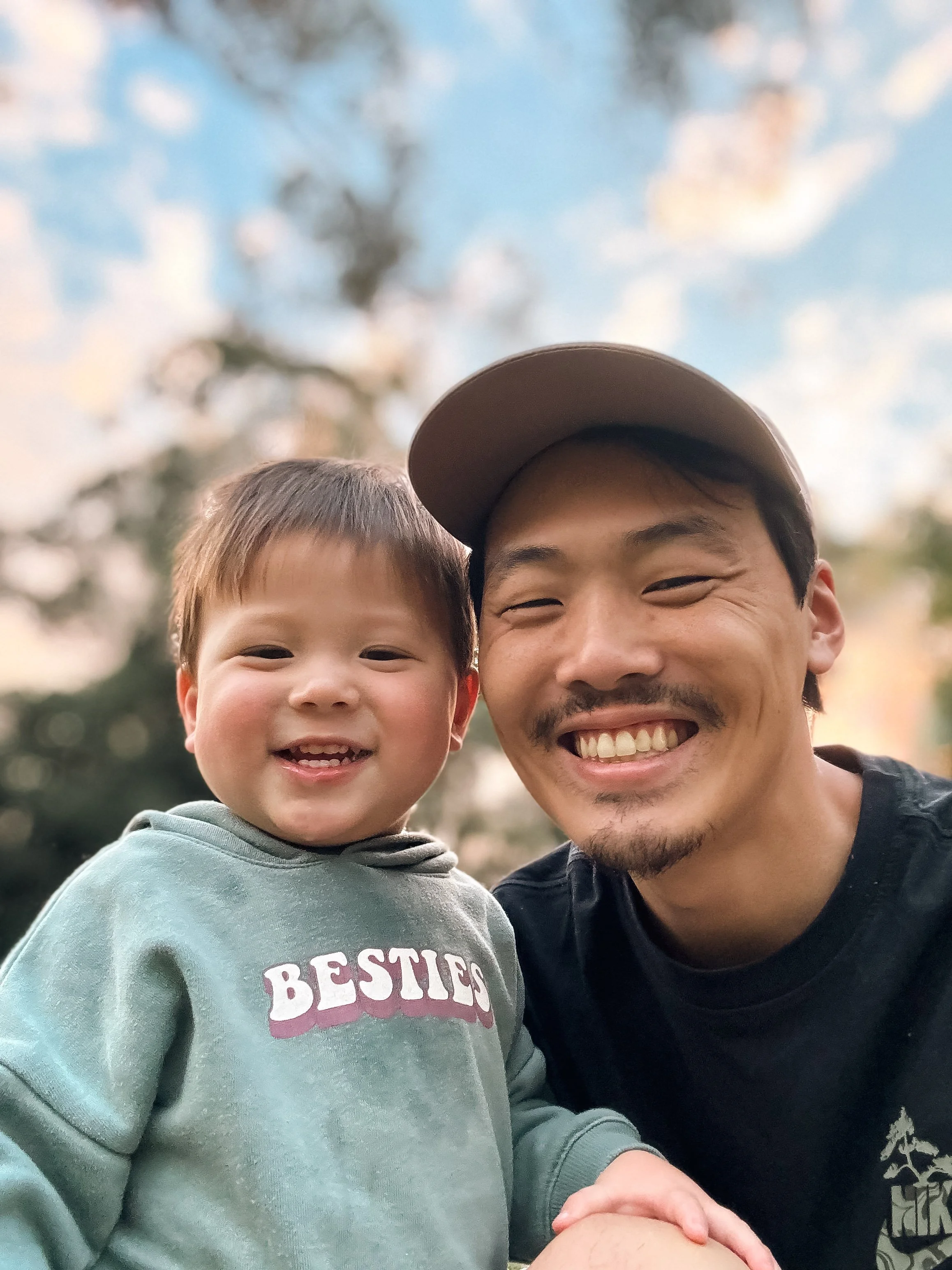 Close-up of a smiling man and a young boy outdoors, with a blue sky with clouds and blurred trees in the background.