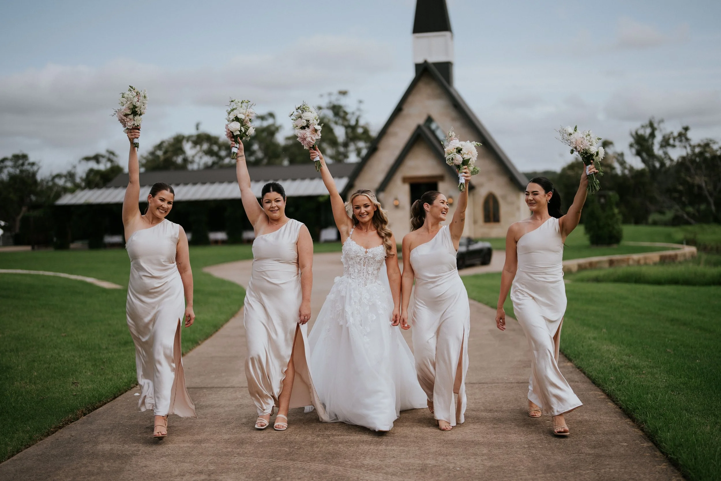 Group of five women, including a bride in a white wedding gown, celebrating outdoors with bouquets of flowers, walking along a path in front of a church.