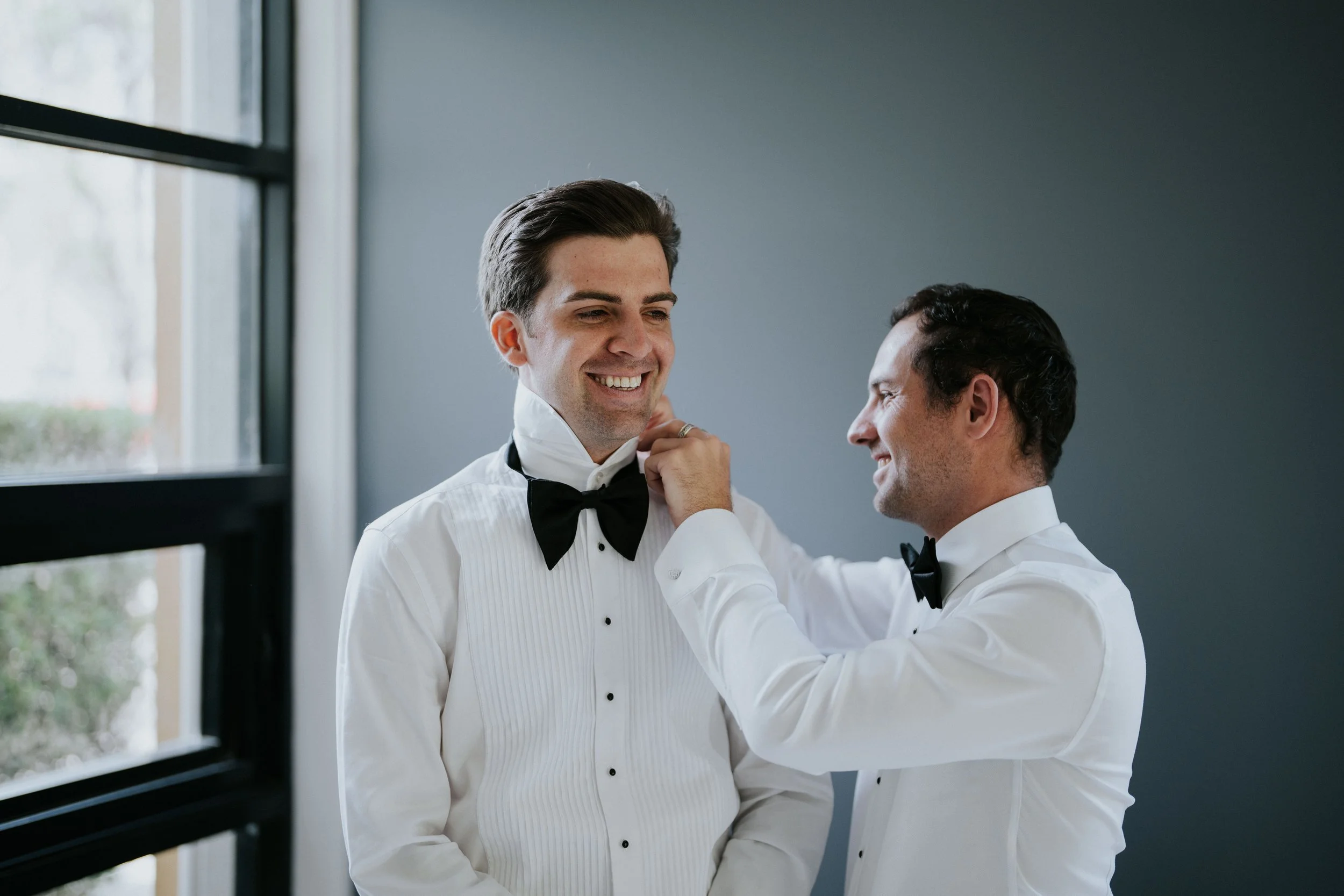 Two men in tuxedos are preparing for a formal event, with one helping the other adjust his bow tie near a window with natural light.