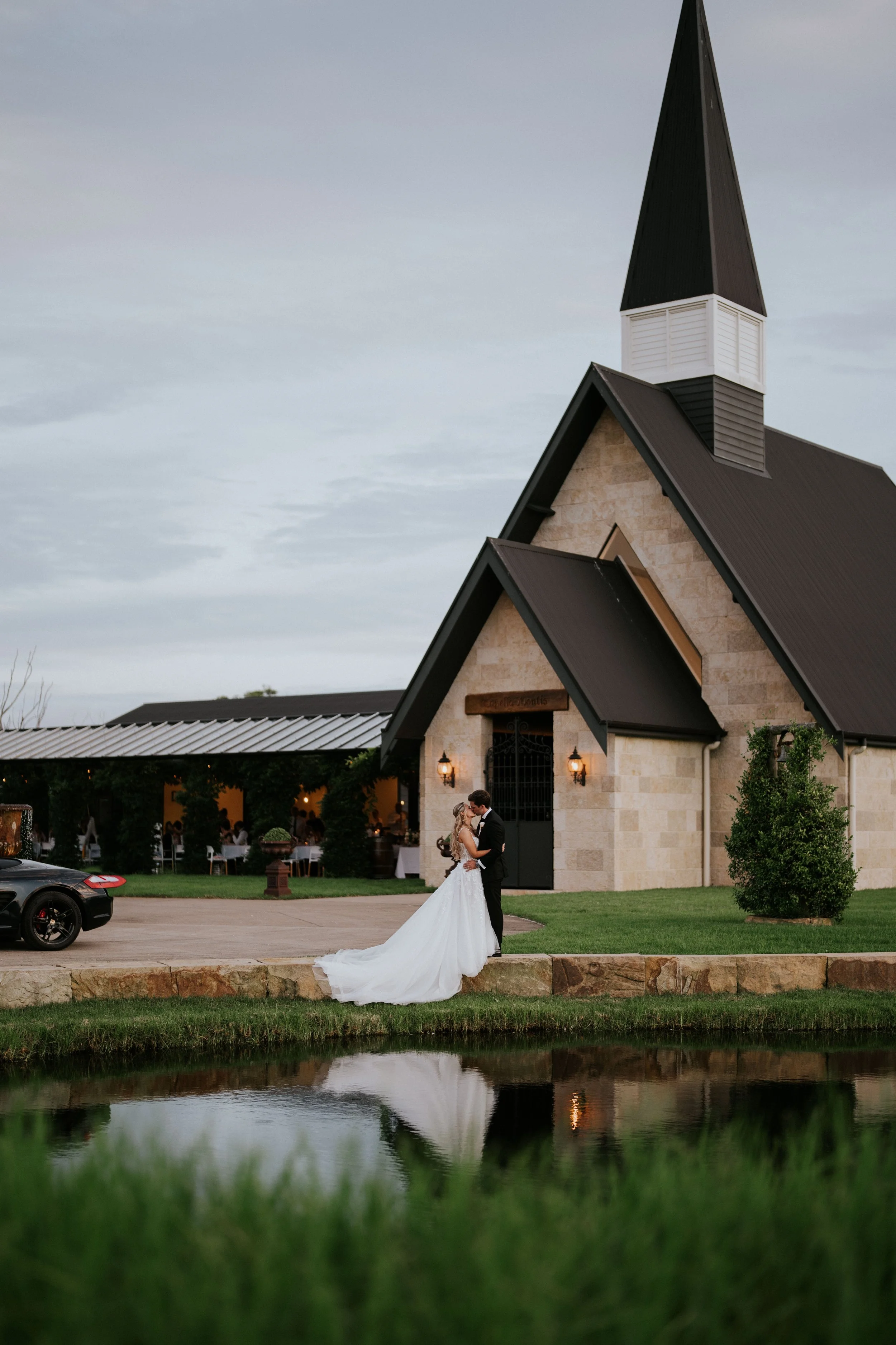 A bride and groom share a dance outside a church with a modern steeple during twilight, reflected in a pond in the foreground.