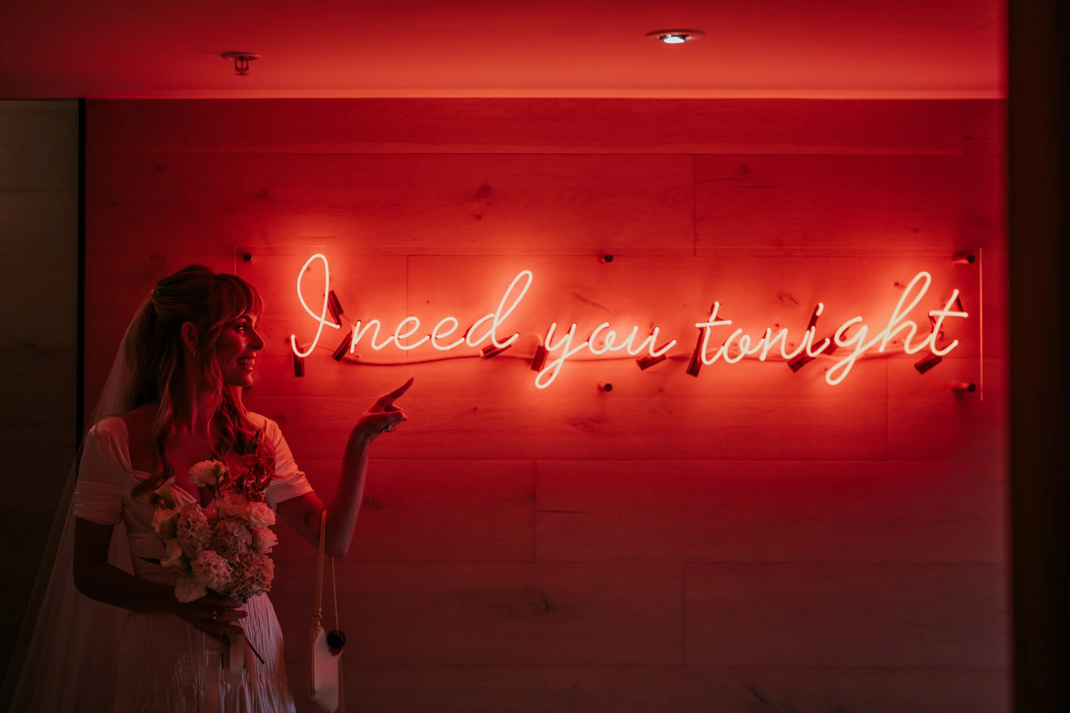 A woman in a wedding dress holding a bouquet of flowers, pointing upward, standing in front of a neon sign that reads 'needed you tonight' in cursive with red lighting.