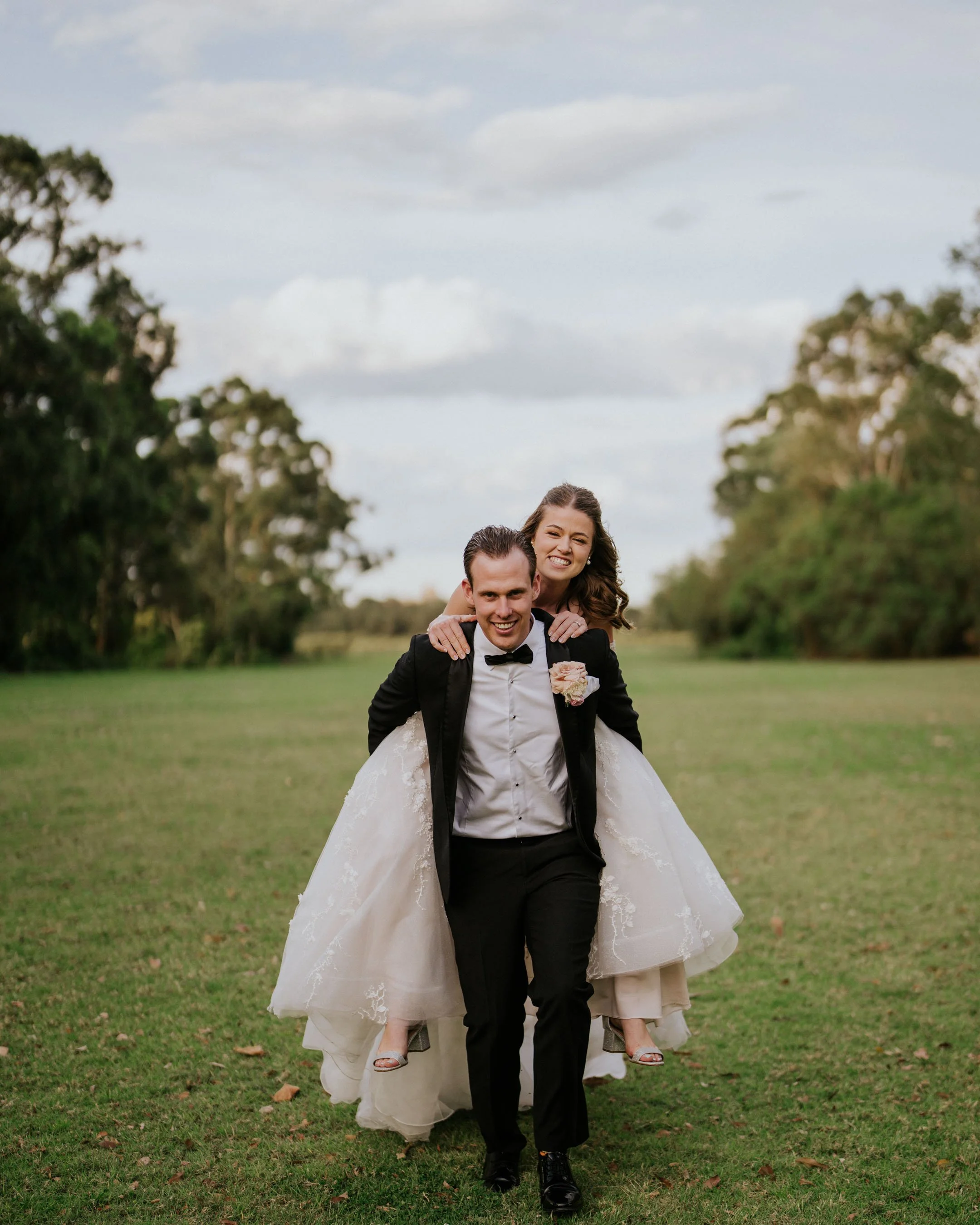 A groom carrying a bride on his back outdoors in a park, both smiling, with trees and a cloudy sky in the background.