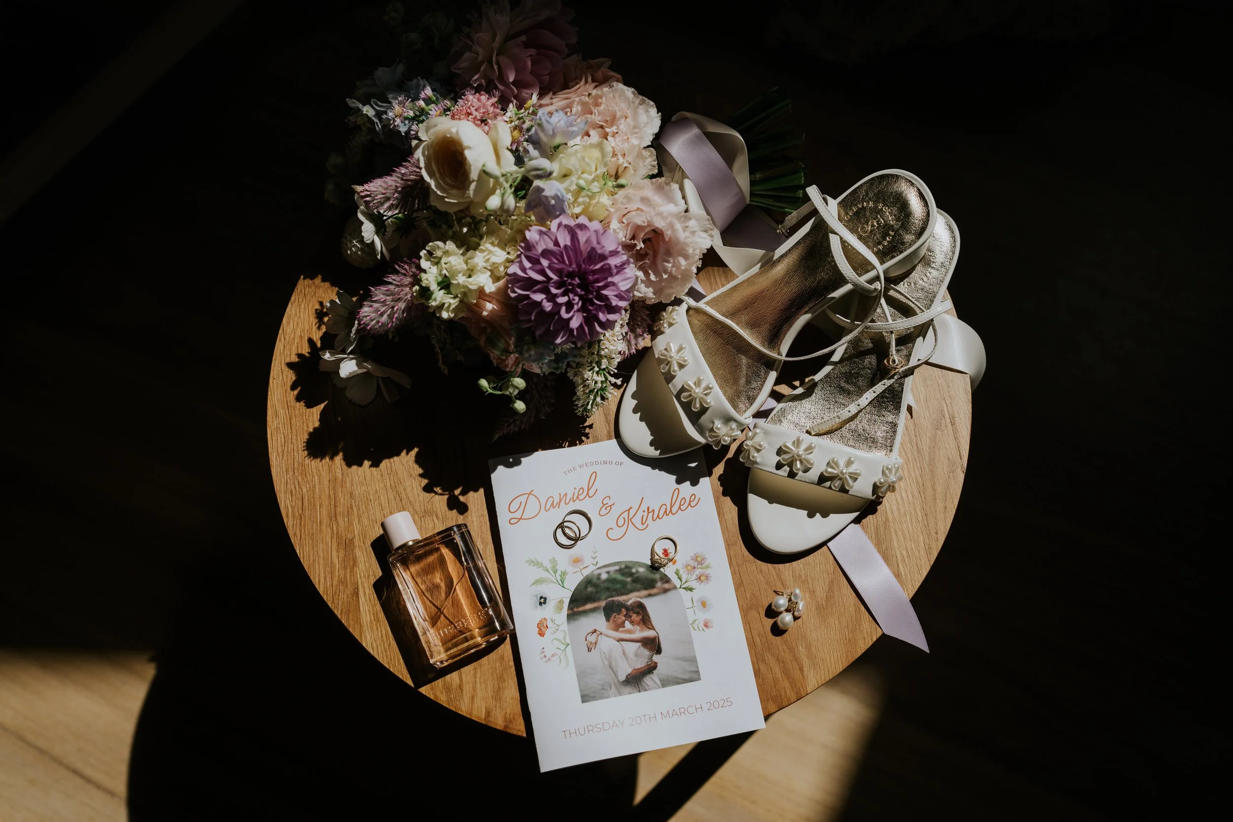 Wedding bouquet with pink and white flowers, white high-heeled shoes decorated with pearls, an invitation card with the names Daniel and Kiralee, a perfume bottle, and wedding rings on a wooden table with dark background lighting.