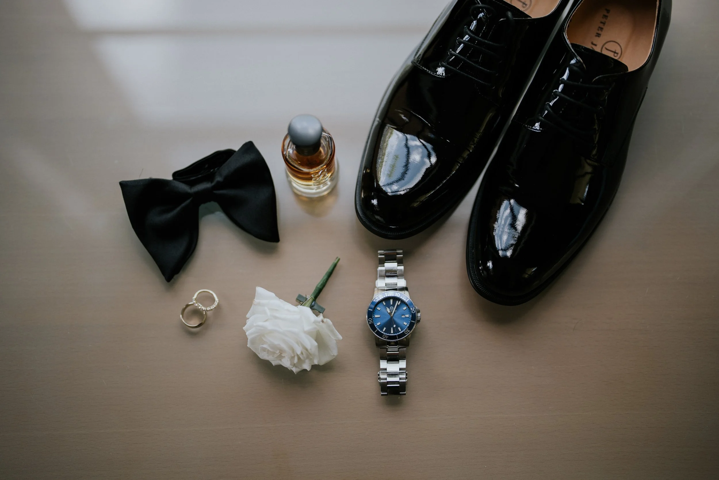 A pair of shiny black dress shoes, a silver wristwatch with a blue face, a crumpled tissue, a gold ring, a pair of gold wedding bands, a bottle of cologne, a black bow tie, a green question mark, and a silver bracelet are arranged on a light-colored 