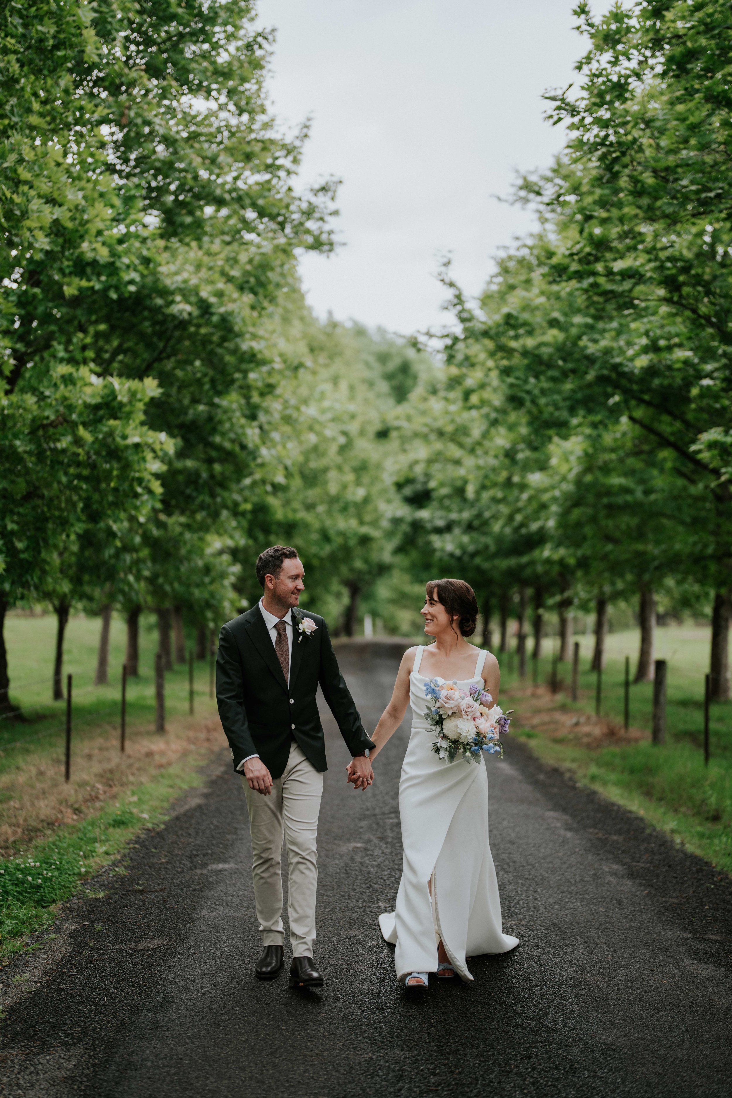 A happy bride and groom walk hand in hand down a tree-lined path, smiling at each other. The bride wears a white wedding gown and holds a bouquet of flowers, while the groom is dressed in a dark suit with a boutonniere. The scene is outdoors with lush green trees on both sides of the path.