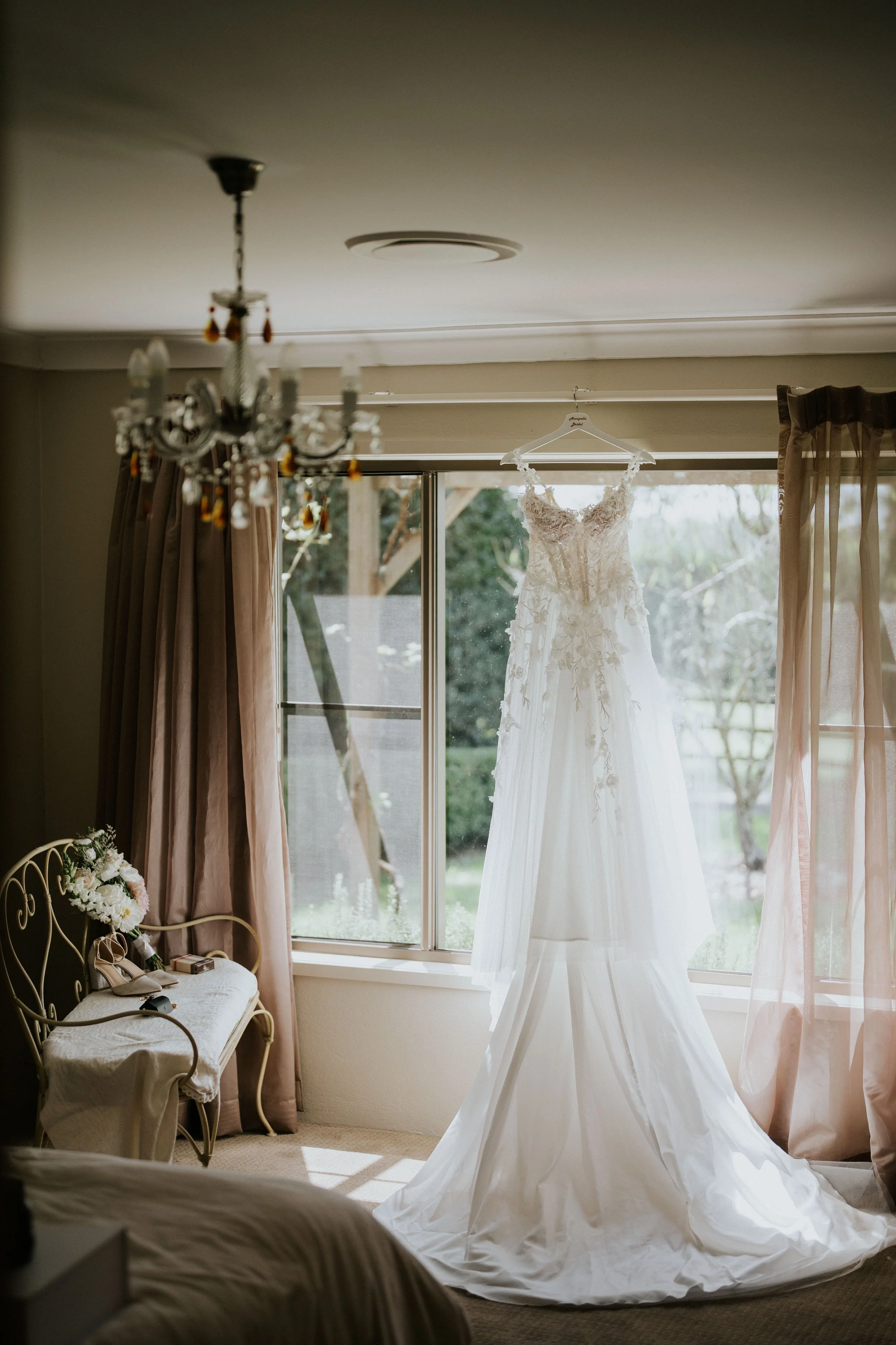 A wedding dress hanging on a hanger in front of a window with curtains, with sunlight illuminating it in a bedroom.
