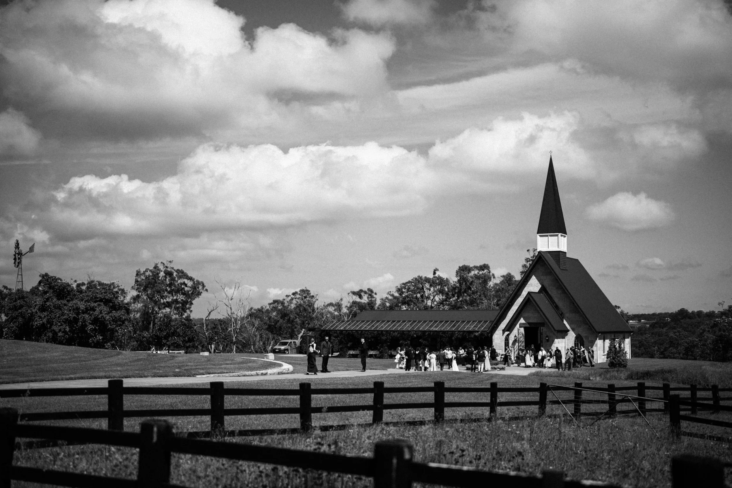 A black and white photo of a church with a tall steeple, surrounded by a crowd of people gathered outside, with trees and a cloudy sky in the background.