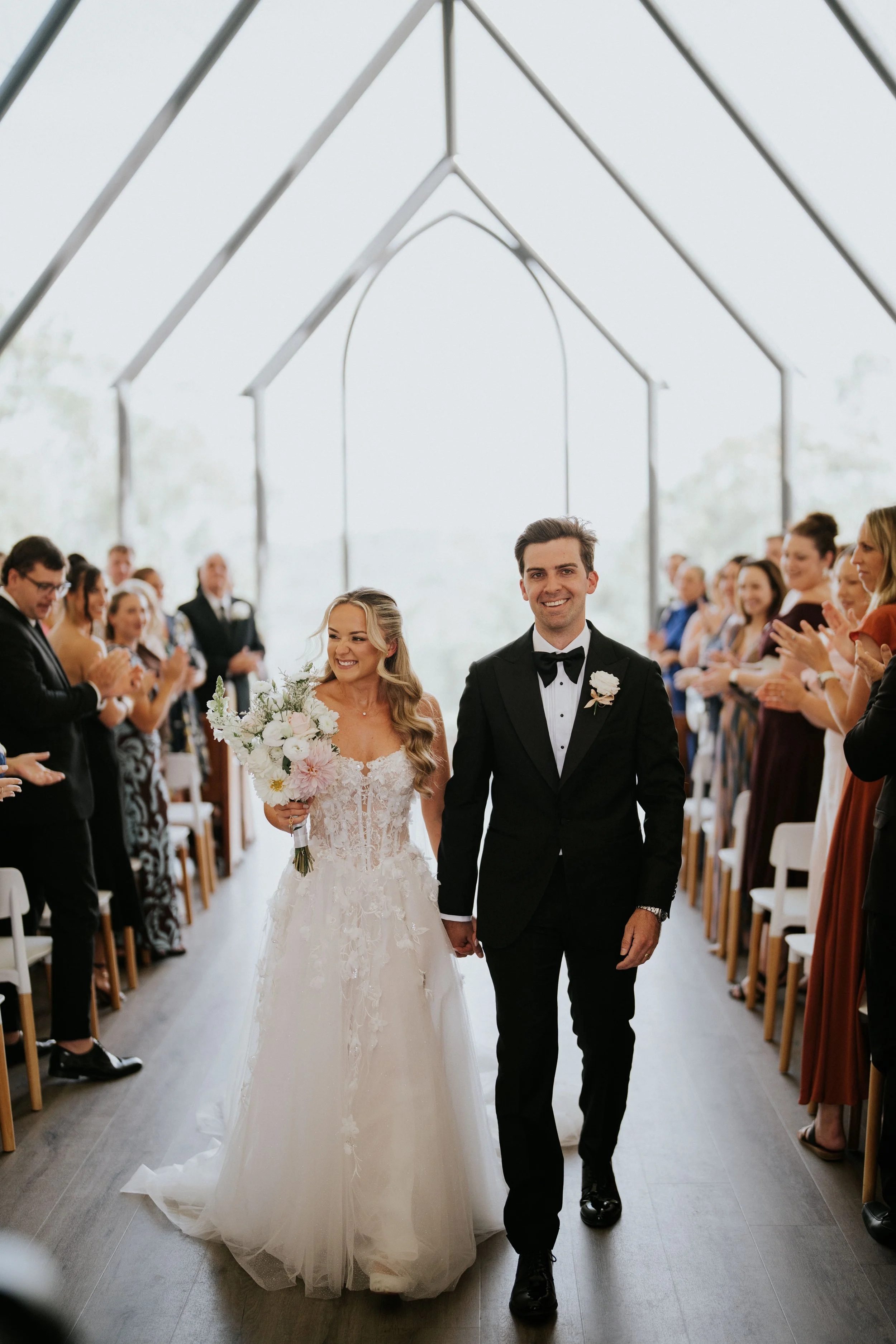 A bride and groom walking down the aisle during their wedding ceremony, surrounded by seated guests clapping and smiling, inside a bright, modern chapel with large glass windows.