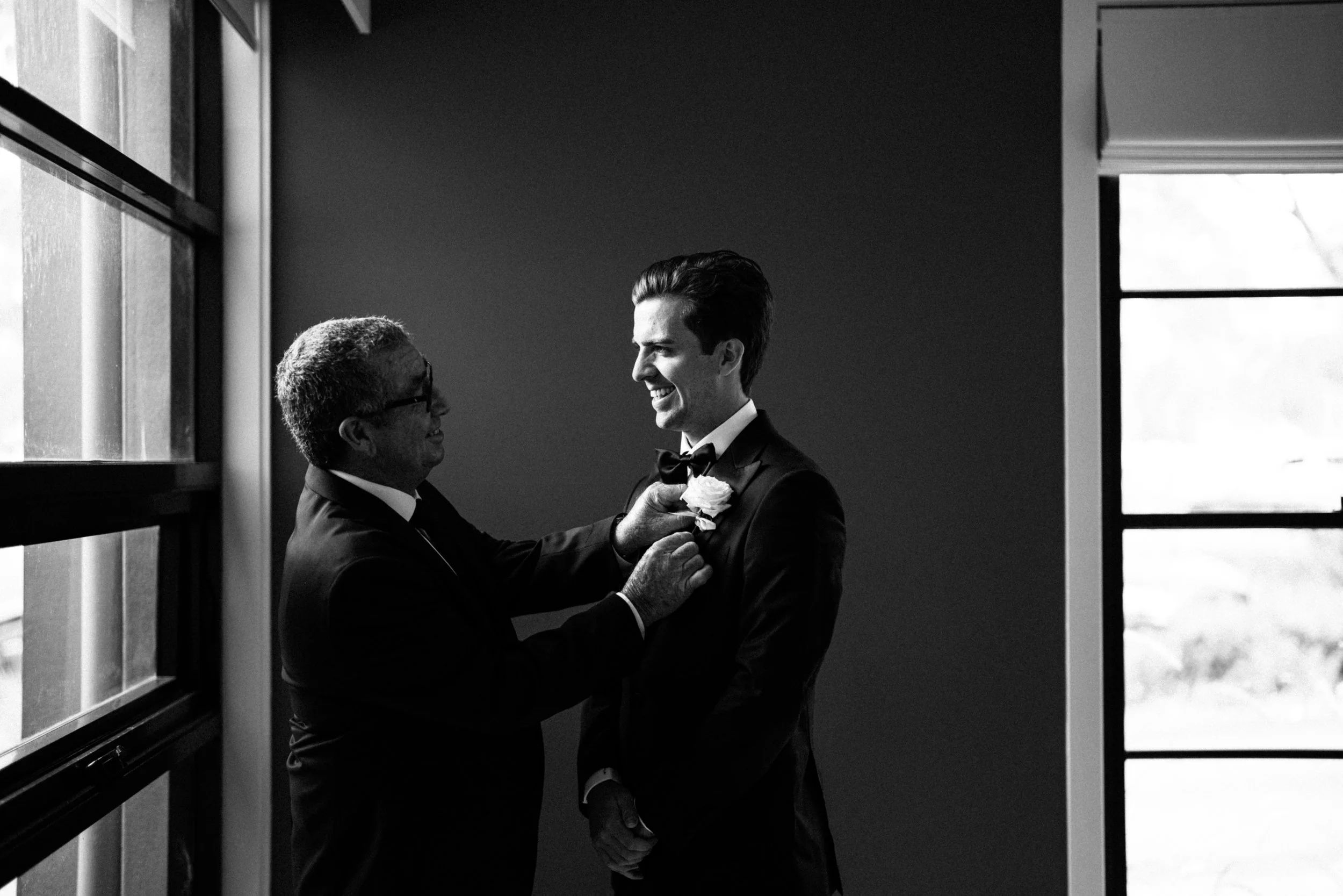 A black and white photo of two men in suits, smiling at each other, with one adjusting a boutonniere on the other's lapel. They are standing near a window with natural light coming in.