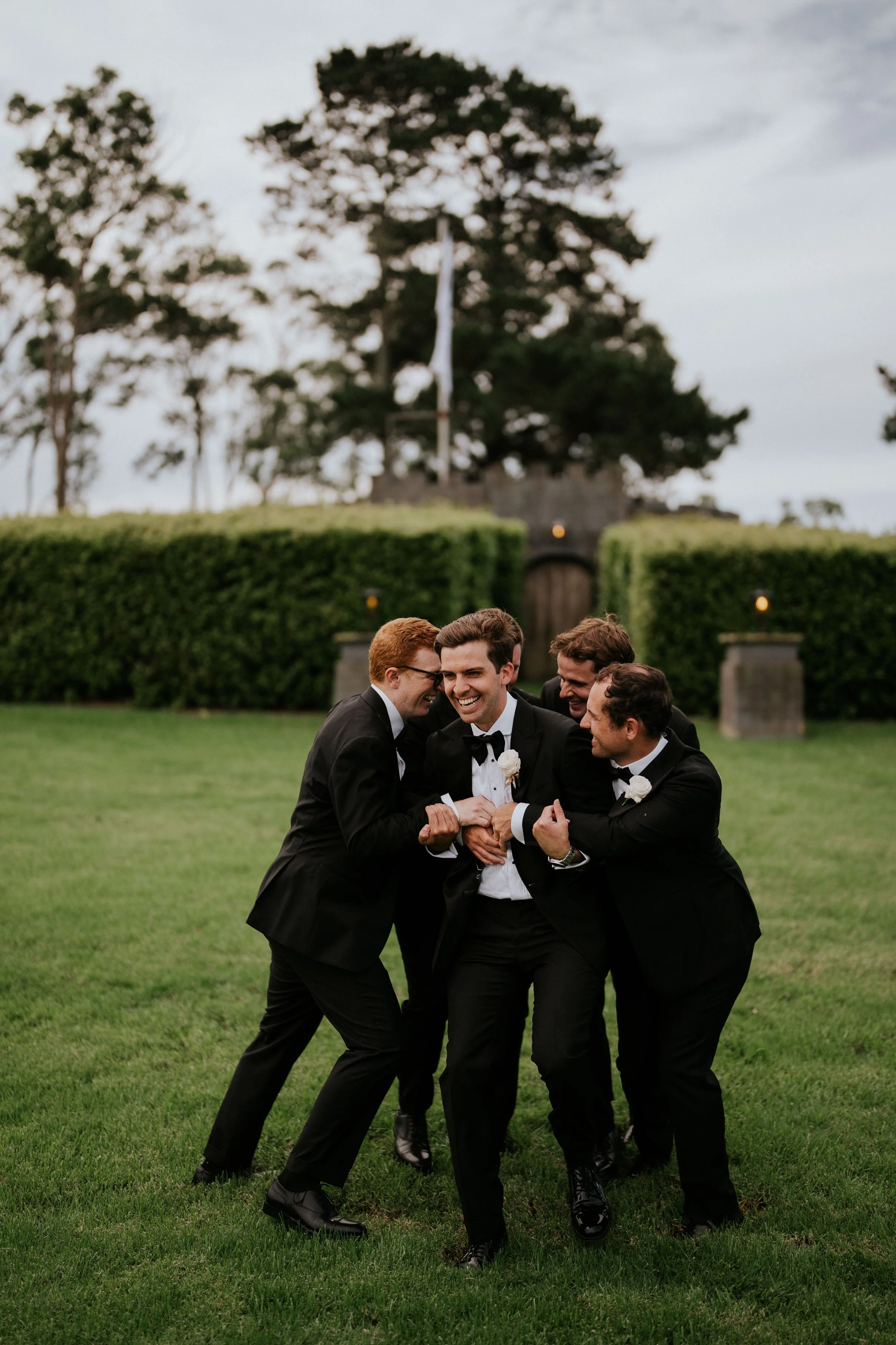 A group of five men dressed in tuxedos laughing and having fun outdoors on a lawn with trees and a hedge in the background.
