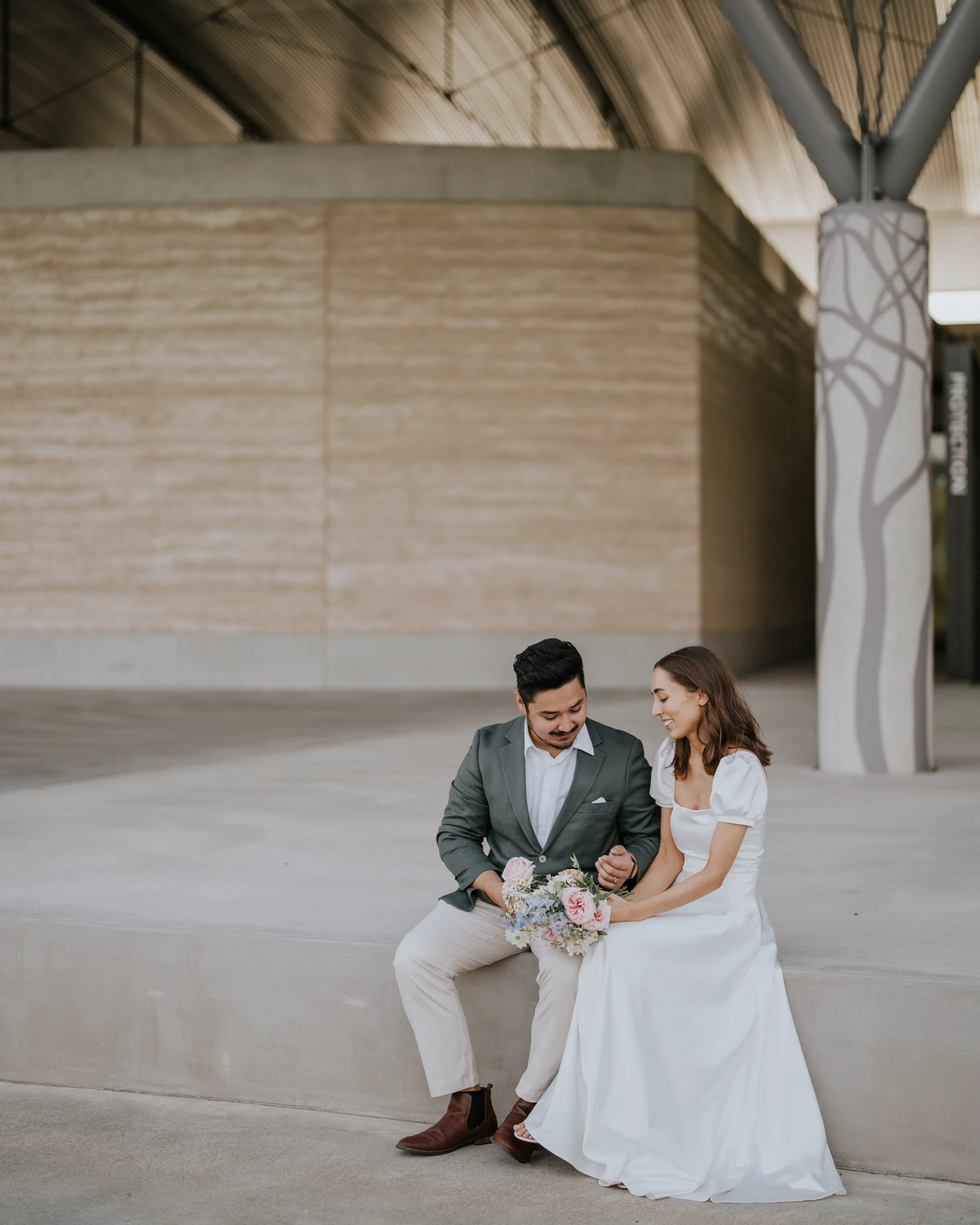 A man and a woman sitting together on a concrete ledge in an outdoor setting, with the man dressed in a suit and the woman in a white dress holding a bouquet of flowers.