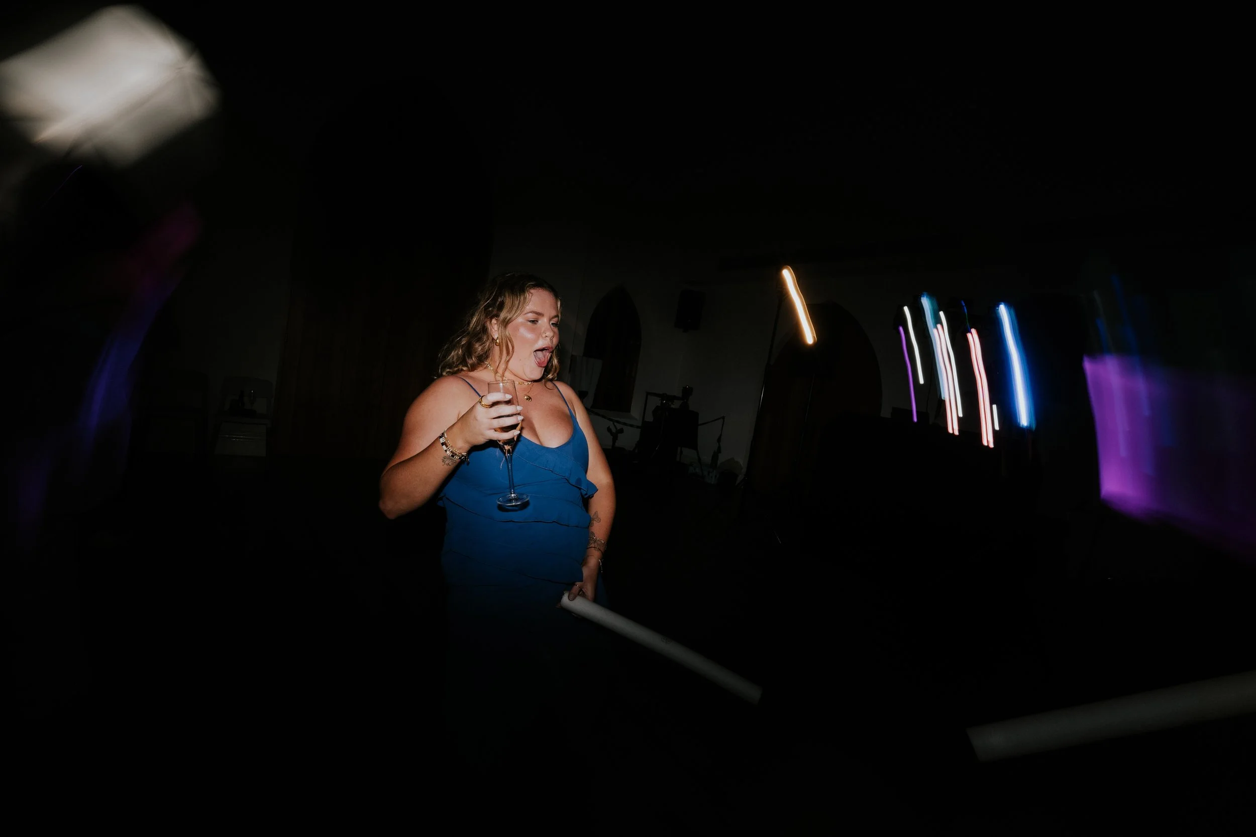 A young woman with curly hair wearing a blue dress. She is holding a drink in her right hand and appears to be singing or talking. The background is dark with colorful vertical light streaks, suggesting a night party or club setting.