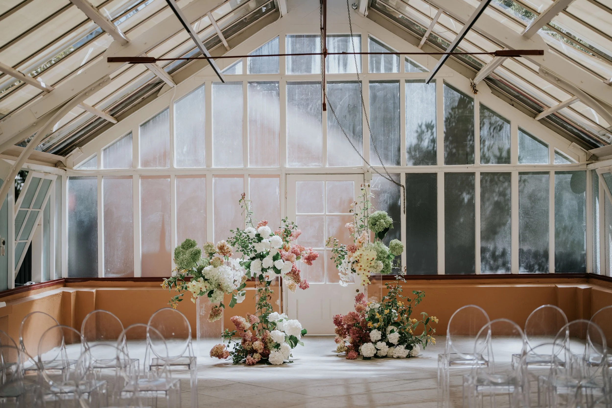 Interior of a conservatory or greenhouse with large windows and a high ceiling, decorated with flower arrangements and transparent chairs, likely set up for a wedding or event.