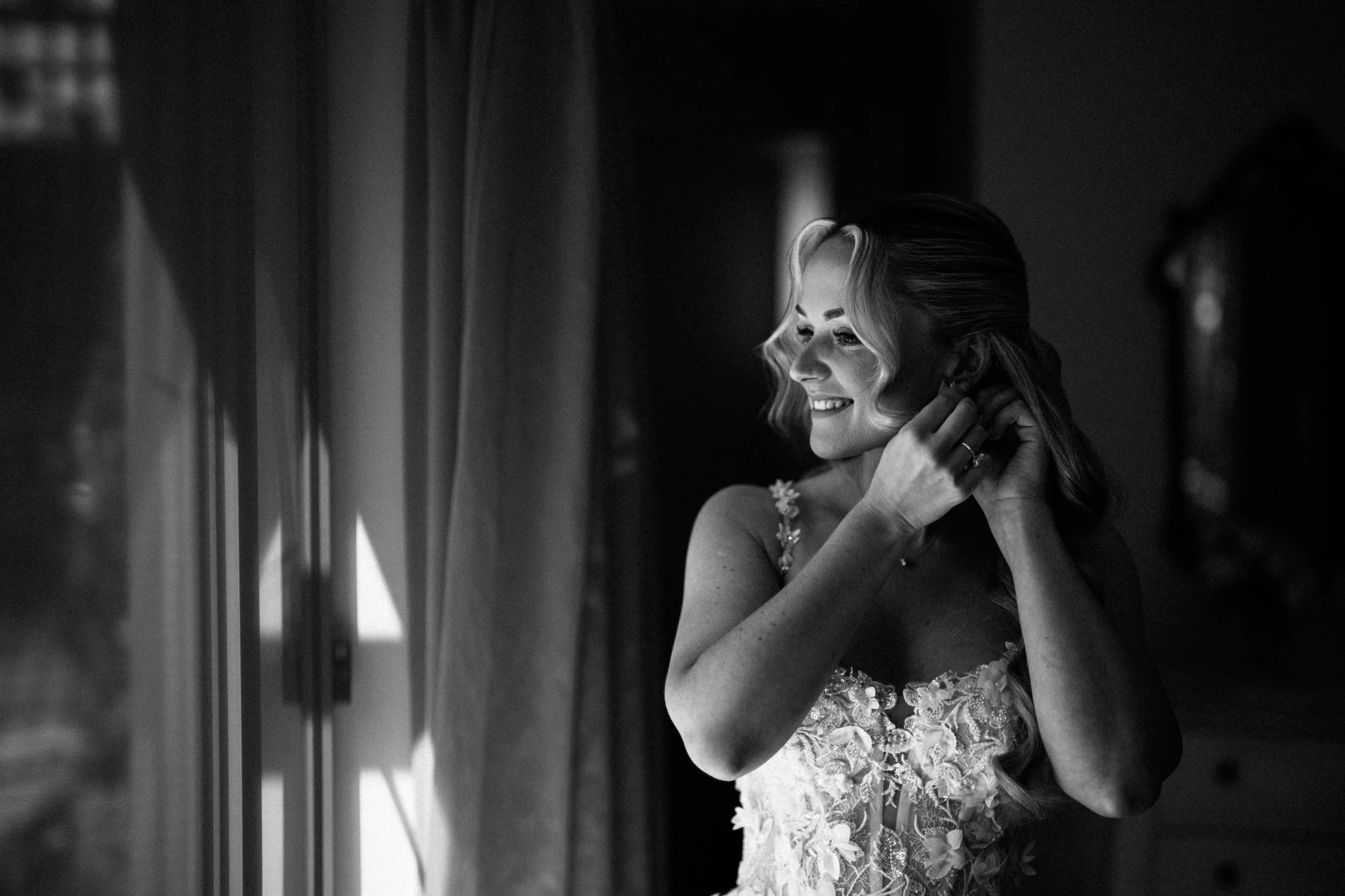 A smiling woman in a wedding dress adjusts her earring while looking out of a window.