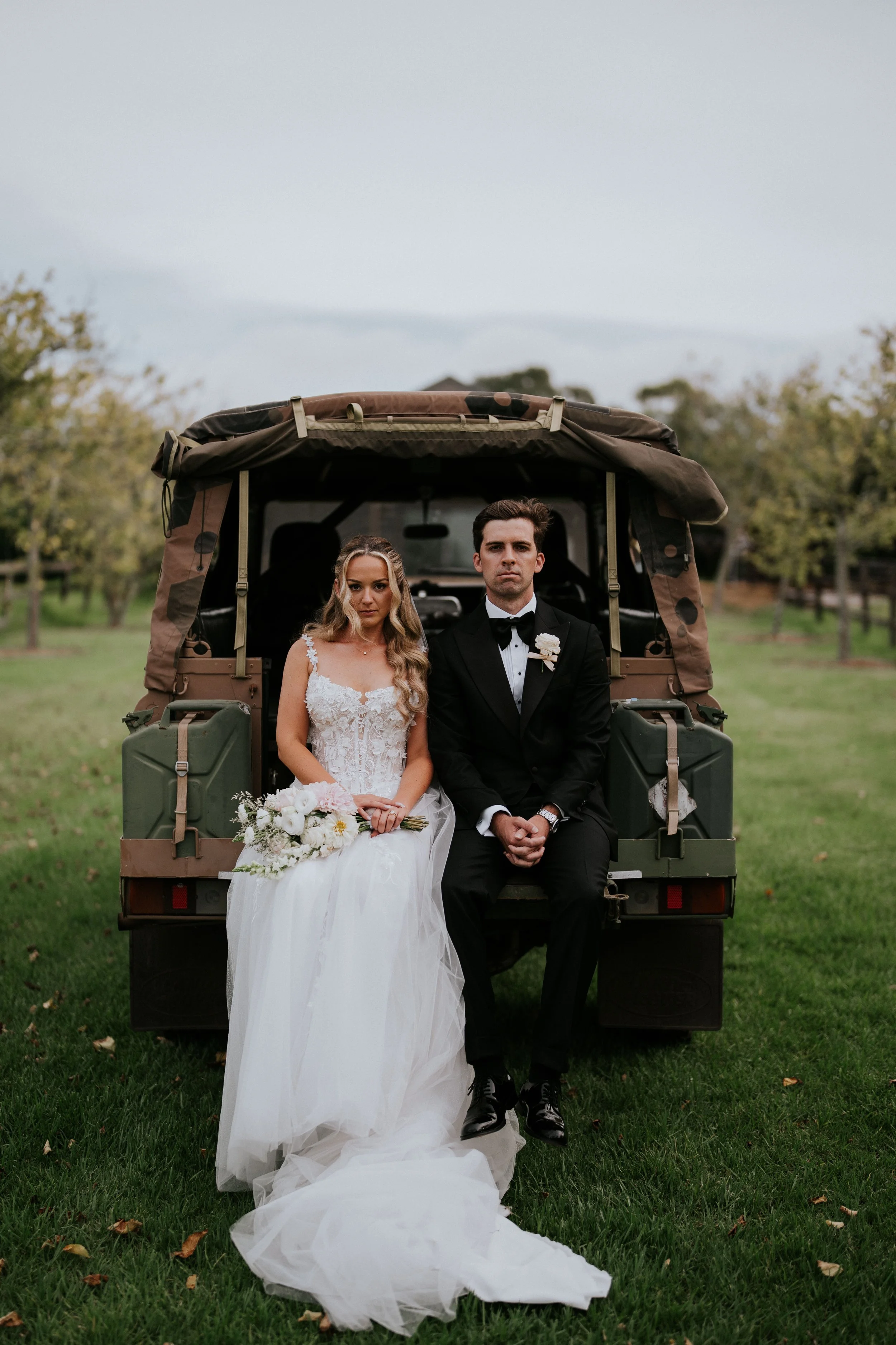 A bride in a white wedding dress holding a bouquet and a groom in a black tuxedo sitting on the back of a military-style truck in an outdoor setting with grass and trees.