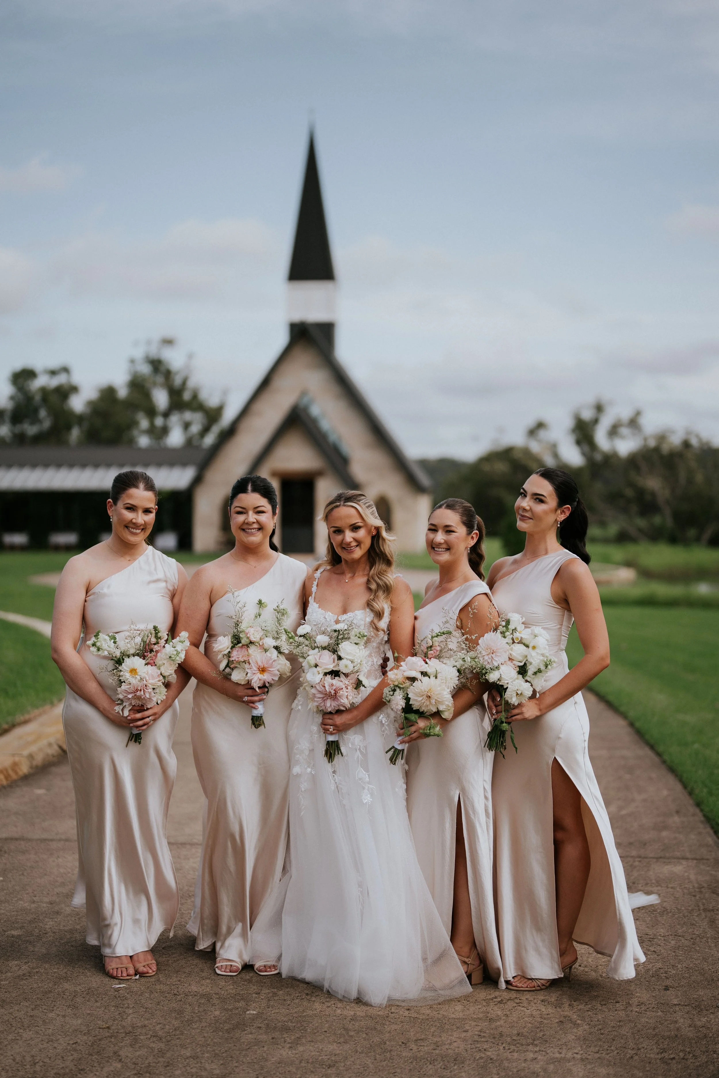 A bride and five bridesmaids in cream-colored dresses holding bouquets of white and pink flowers, standing on a path outside a church with a tall steeple and a lush green landscape in the background.