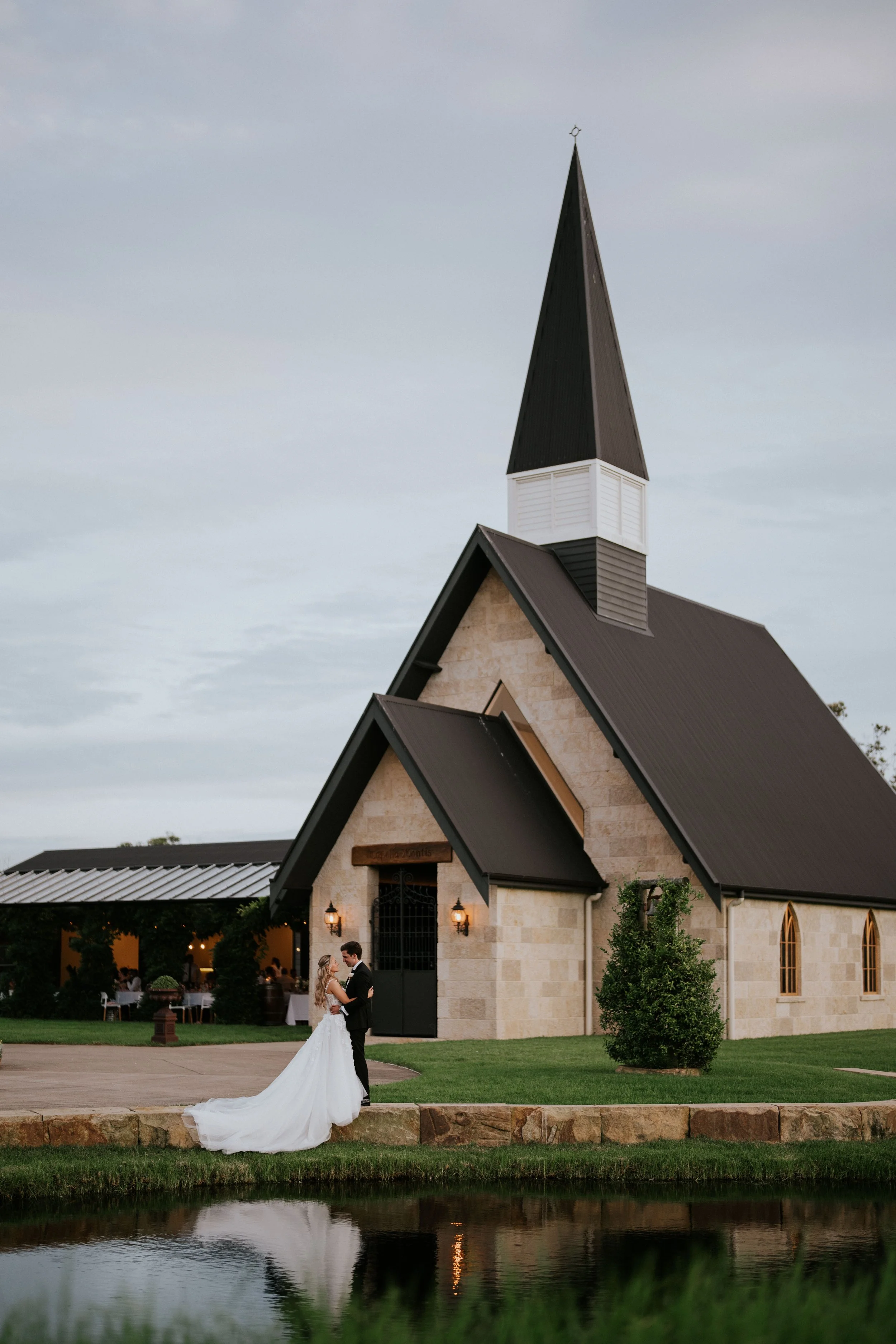 A bride and groom dancing outside a church, with the bride in a white wedding gown and the groom in a black suit. The church features a tall steeple and brick construction, with an outdoor gathering of people in the background.
