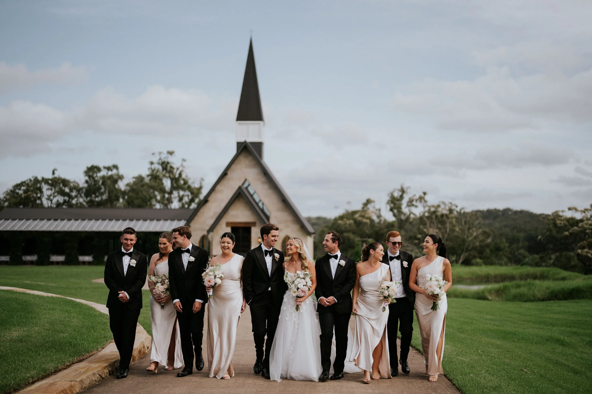 Group of wedding attendees including brides and grooms walking outdoors on a pathway in front of a church, with a steeple and trees in the background.