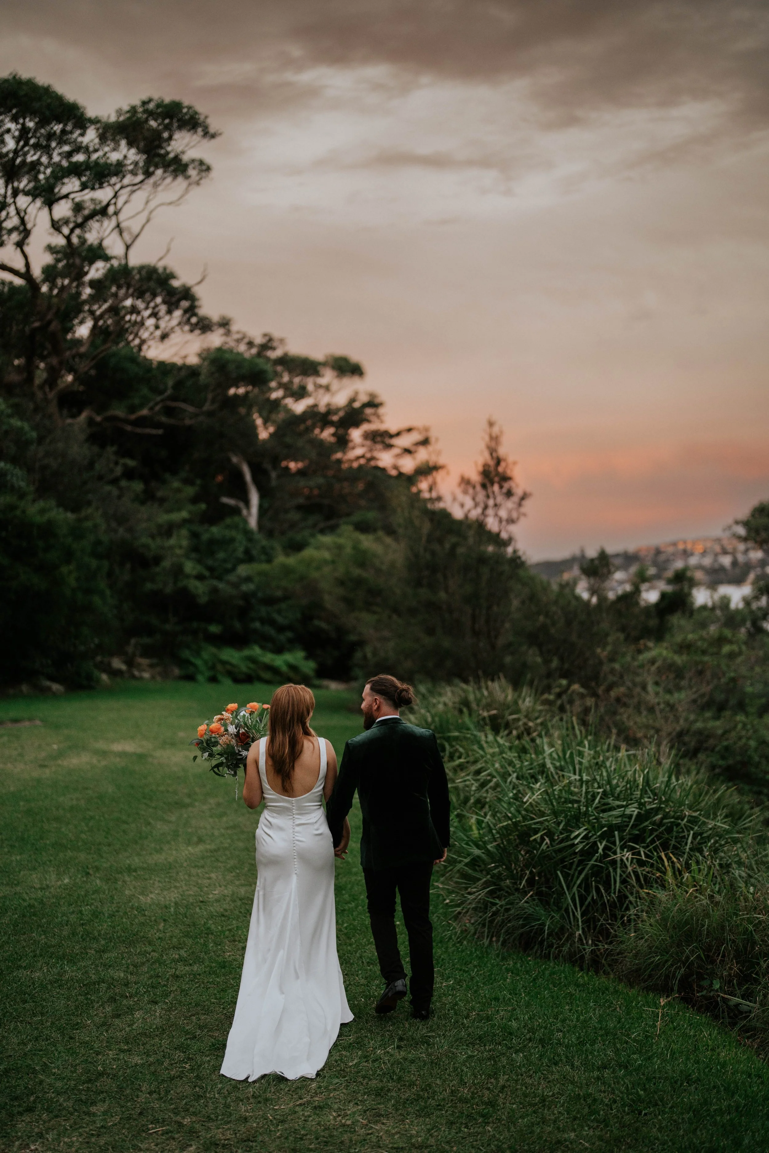 A bride and groom walk hand in hand on a grassy path in a lush garden at sunset. The bride holds a bouquet of flowers, and they are dressed in wedding attire, with trees and a sunset sky in the background.