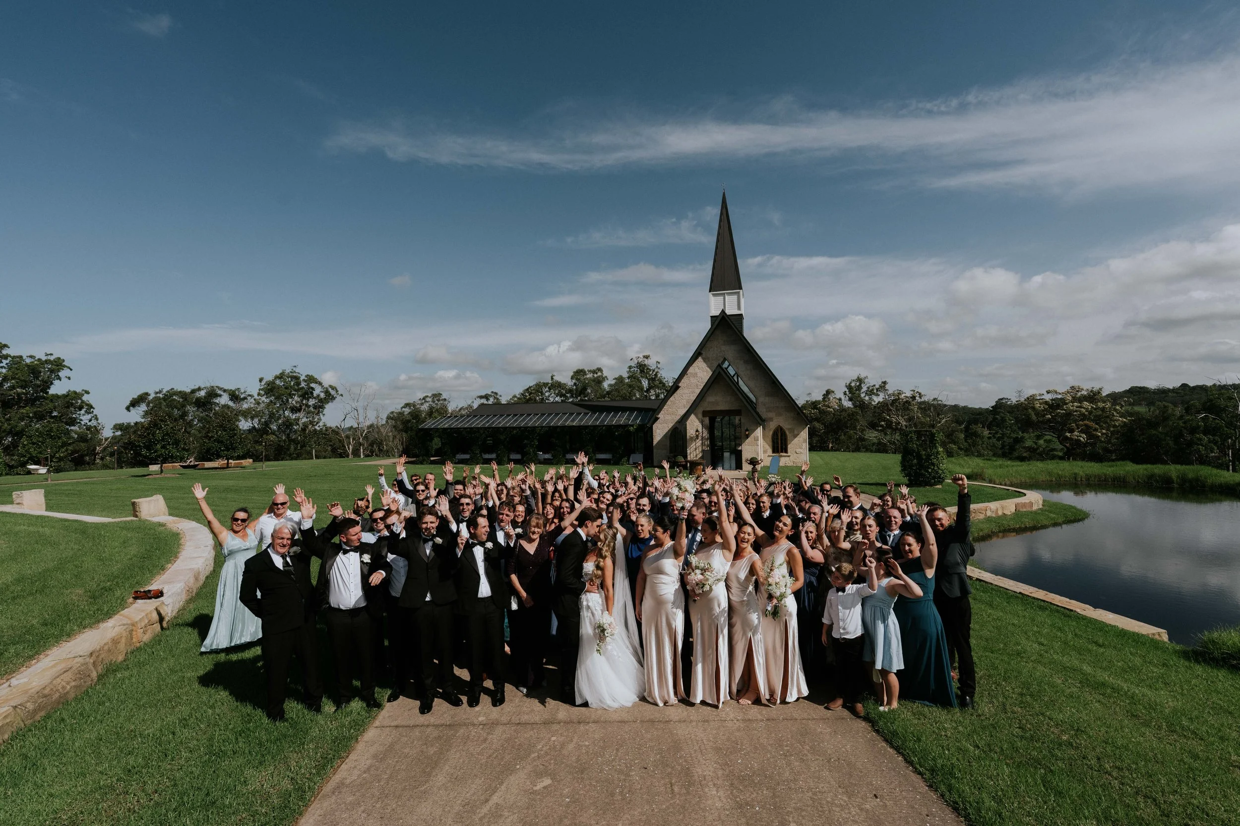 A large group of wedding guests and the bride and groom posing outdoors near a church with a tall steeple, trees, and a small pond under a partly cloudy sky.