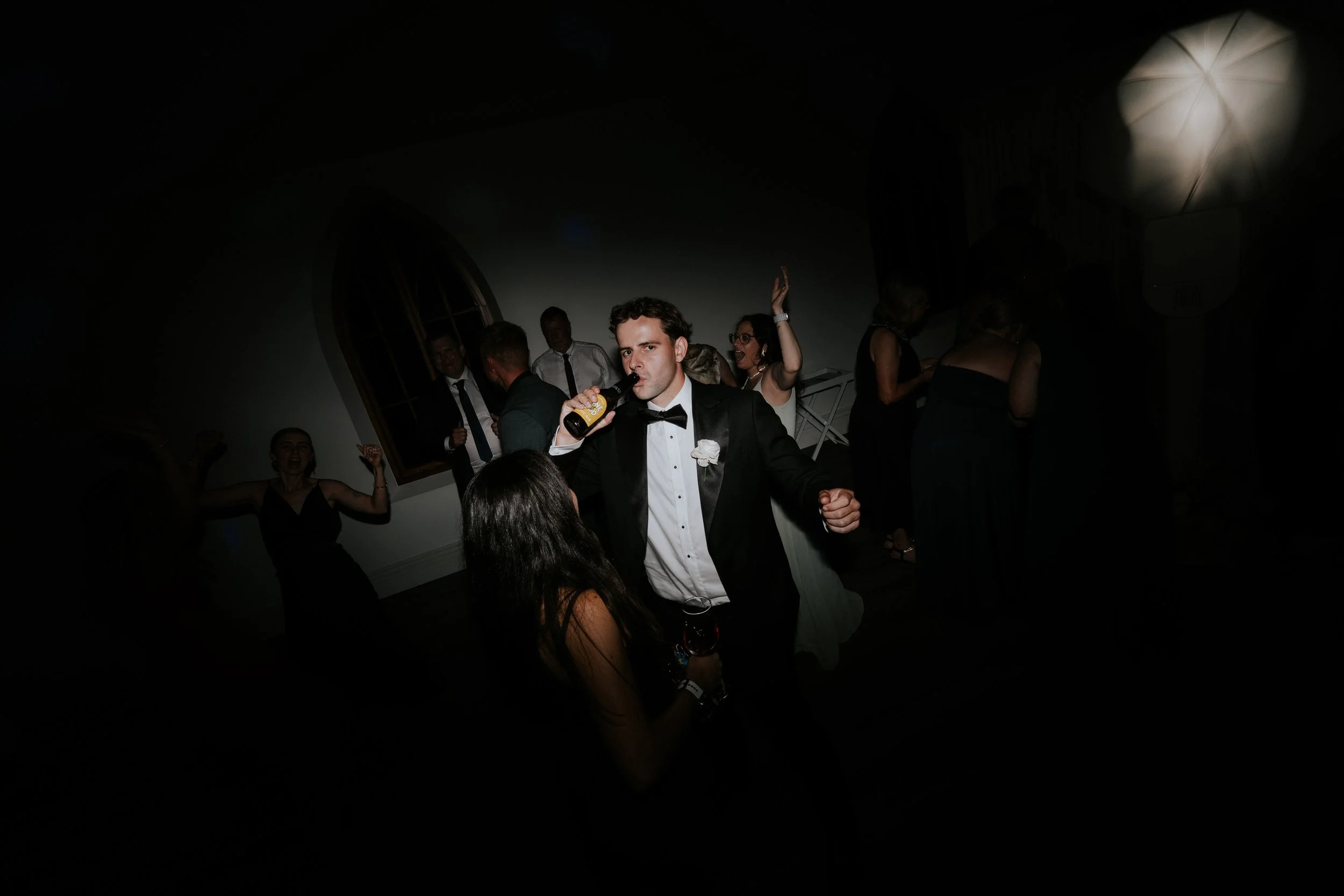 A group of people dancing and having fun at a party or celebration in a dimly lit room. A man in a tuxedo is drinking from a bottle in the foreground, while women and men are dancing and socializing in the background.