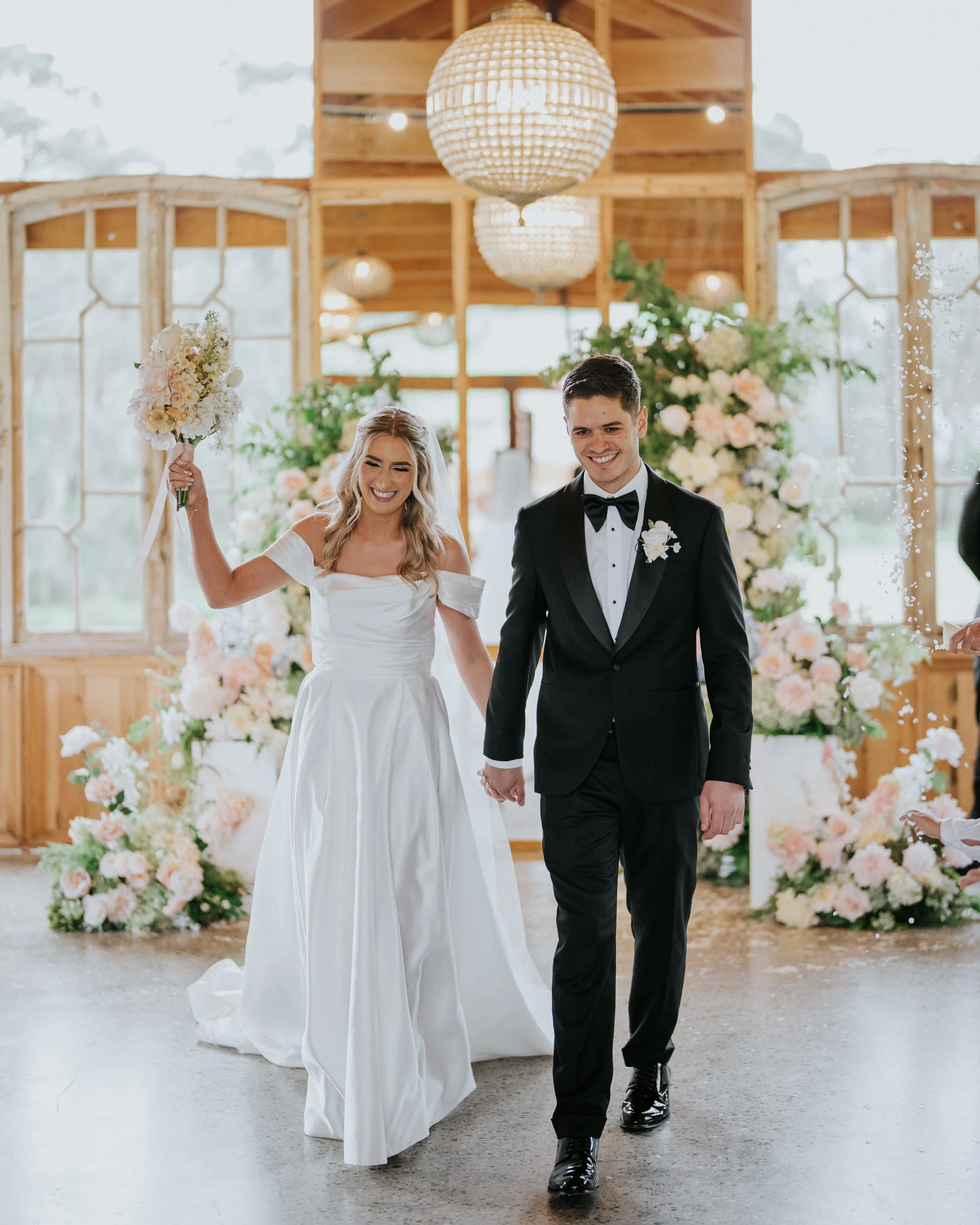 A bride and groom walking hand-in-hand down the aisle during their wedding ceremony. The bride is holding a bouquet of pink and white flowers, and they are smiling happily. The background features floral arrangements and wooden frames with hanging spherical lights.