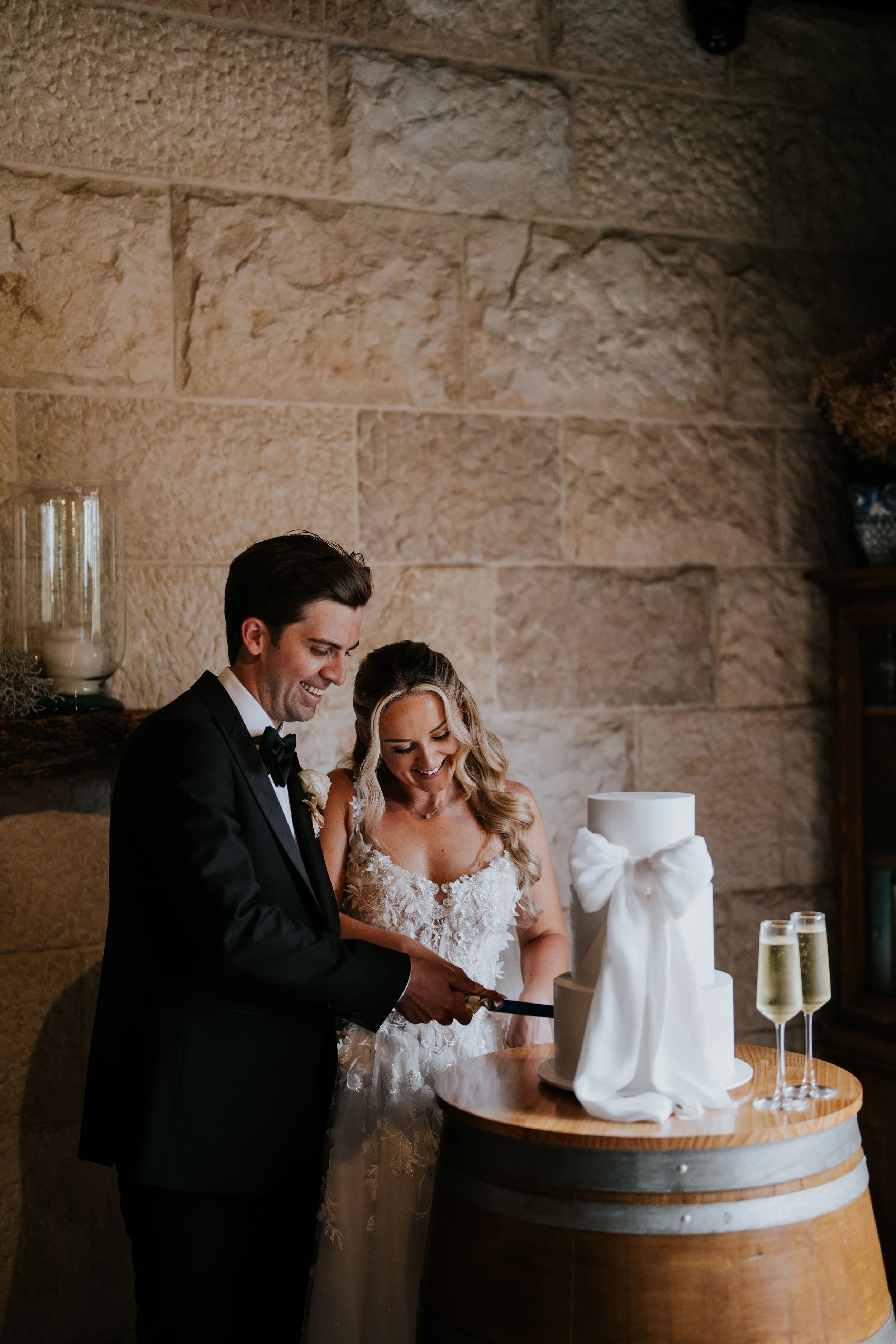 A wedding couple, a man in a tuxedo and a woman in a wedding dress, cutting a wedding cake at a reception with a stone wall background, champagne glasses on a wooden table, and a large white decorative bow on the cake.