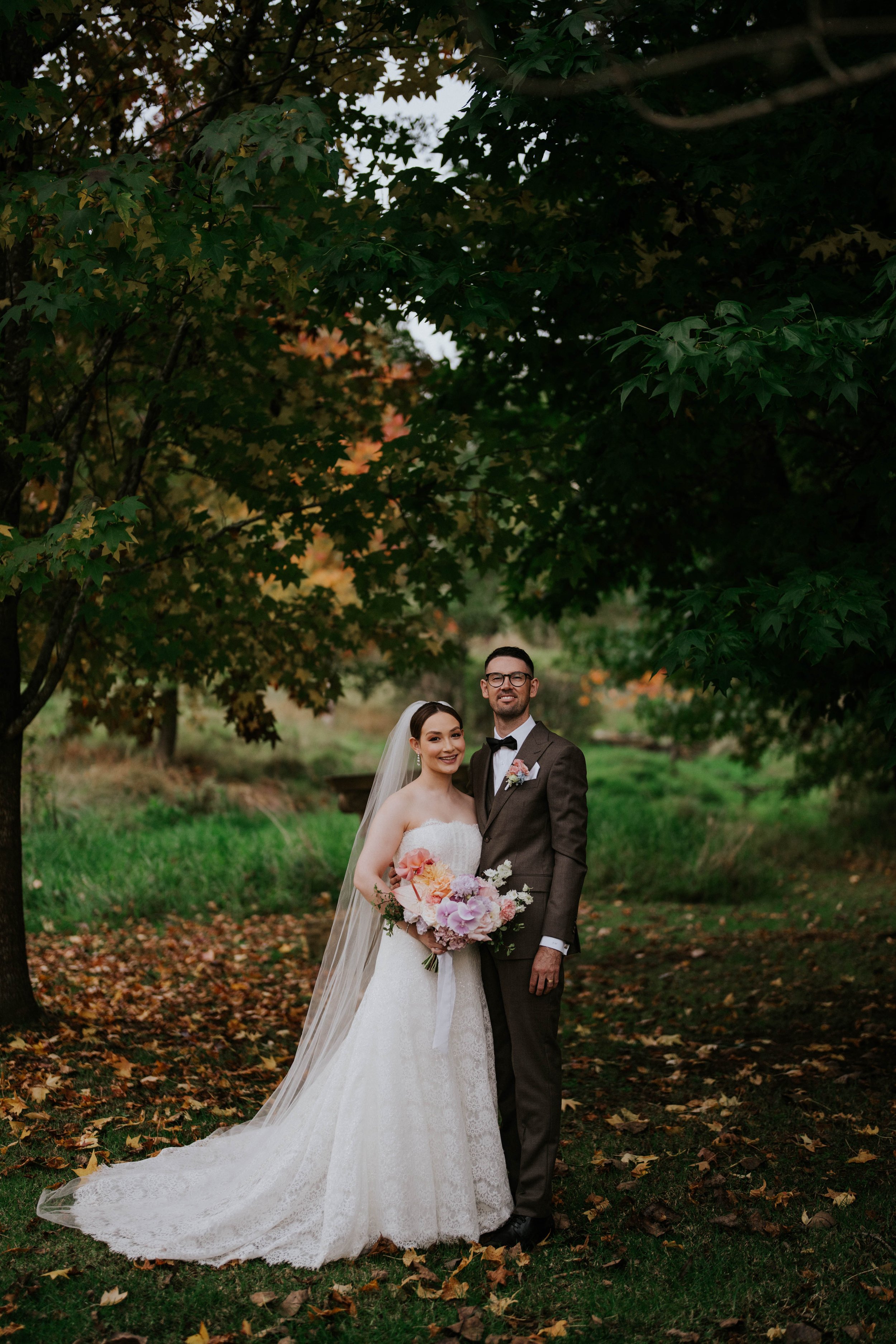 A bride and groom standing in a park with fallen leaves, under a large leafy tree during autumn. The bride is wearing a white wedding gown with a veil and holding a bouquet of pink, lavender, and white flowers. The groom is dressed in a brown suit wi