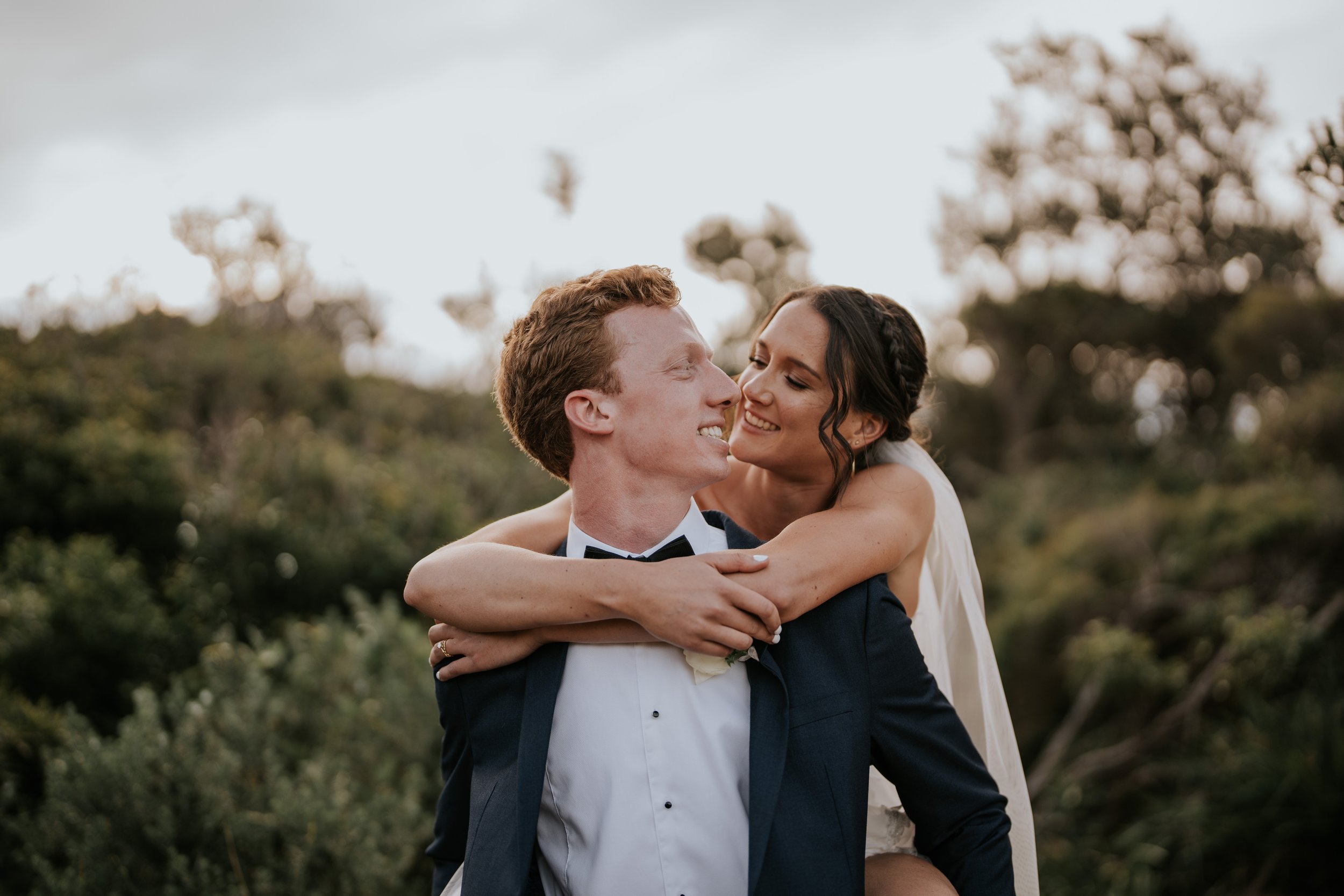 A couple dressed in wedding attire, the woman is hugging the man from behind while they smile at each other outdoors with blurred trees in the background.
