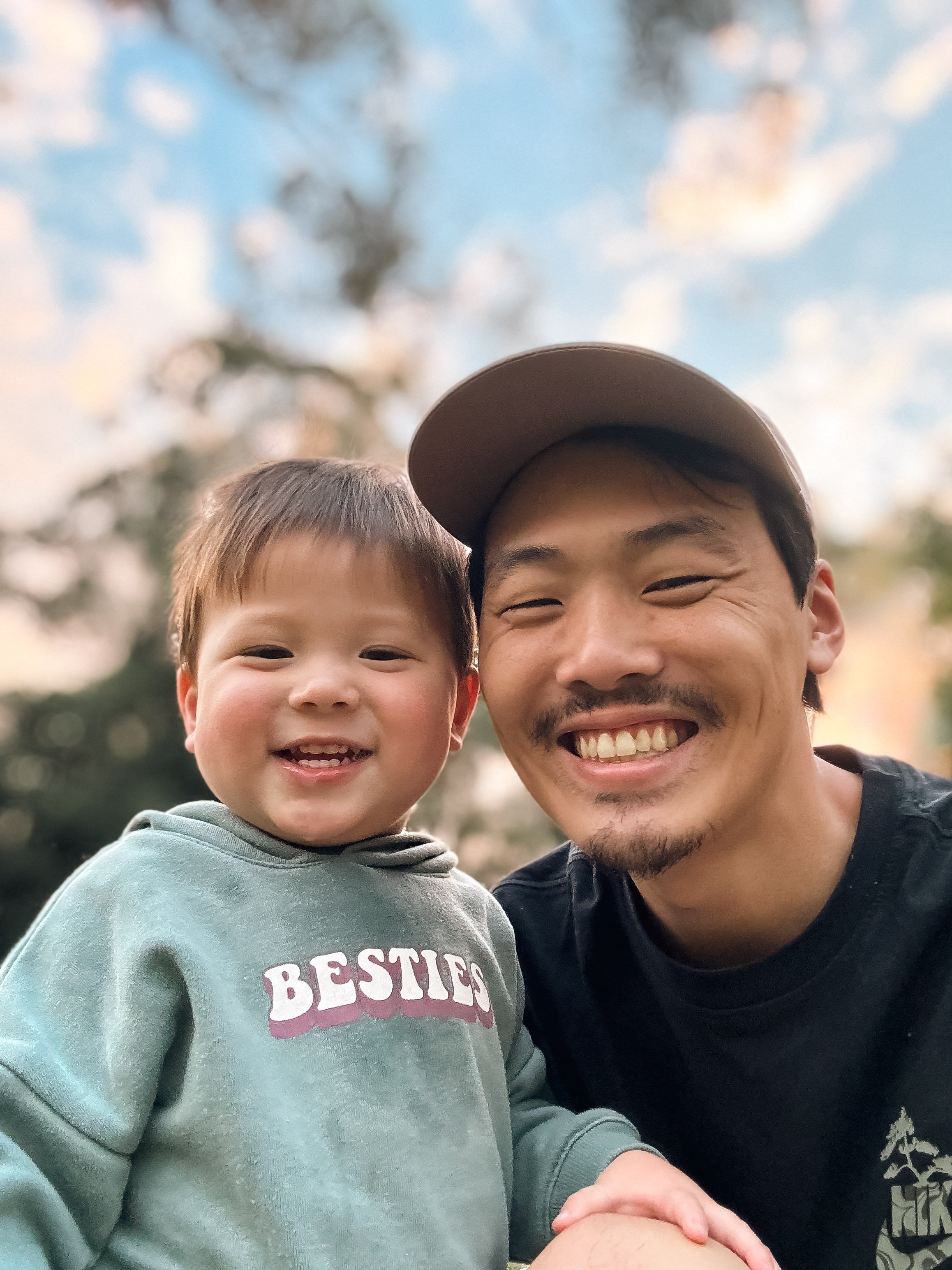 A smiling man with a beard and a cap next to a young boy with short hair, both smiling and holding hands outdoors with a blurred background of trees and sky.
