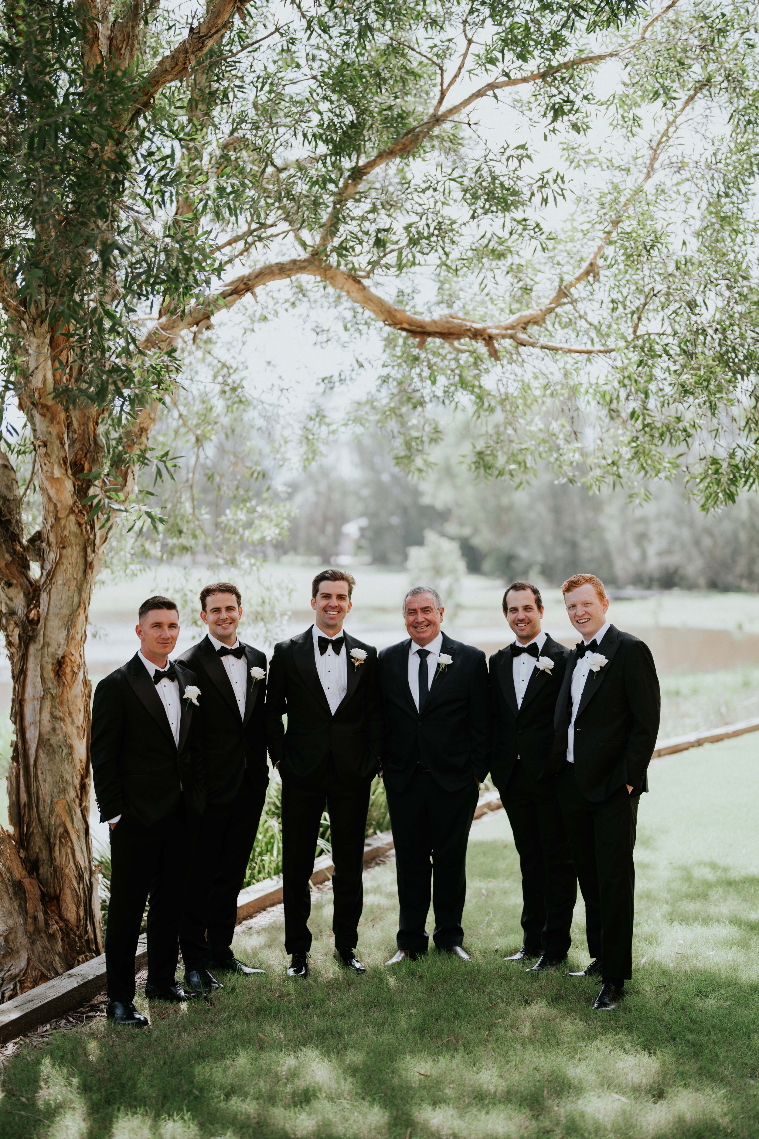 A group of seven men in tuxedos standing outdoors under a large tree, smiling for the camera.