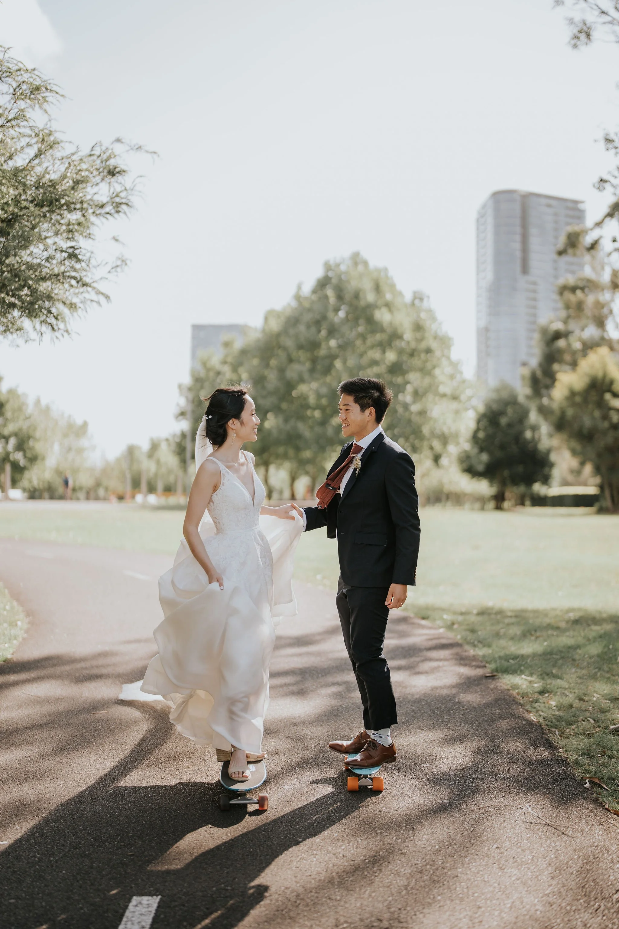 A bride and groom in wedding attire riding skateboards on a park path, holding hands and smiling at each other, surrounded by trees with a cityscape in the background.