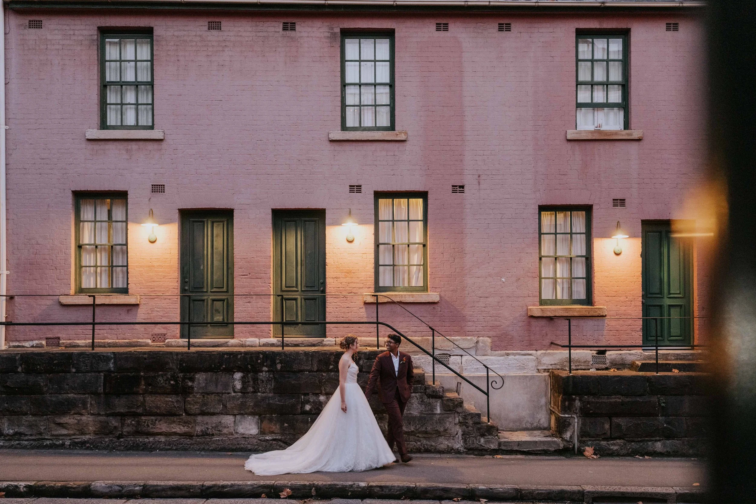 A bride and groom walking down steps in front of a pink brick building at dusk. The bride wears a white wedding dress with a long train, and the groom wears a dark suit. The building has six windows with white curtains and two doors, illuminated by w