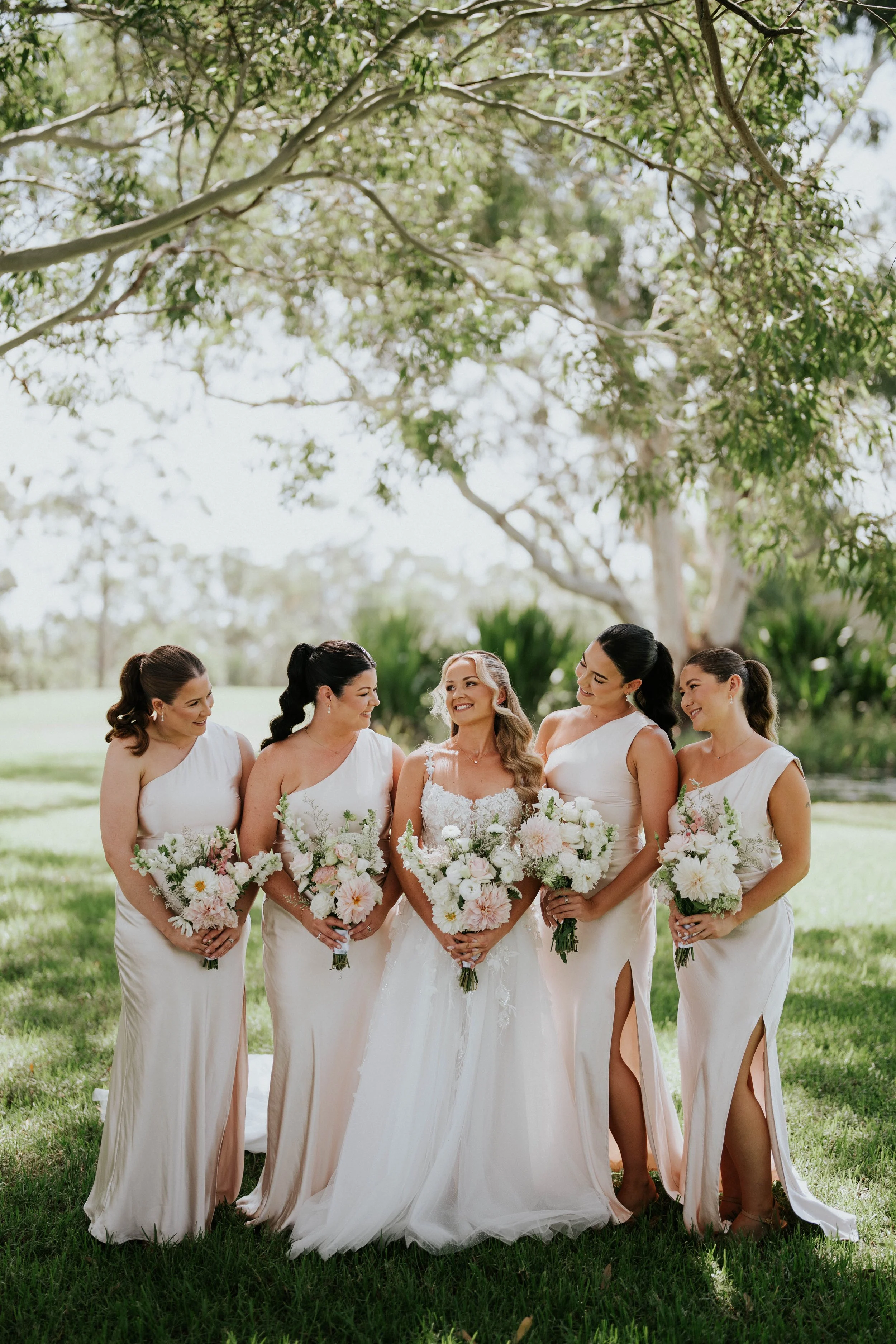 A bride and five bridesmaids standing outdoors on a grassy area, holding bouquets of flowers, with trees and a blue sky in the background.