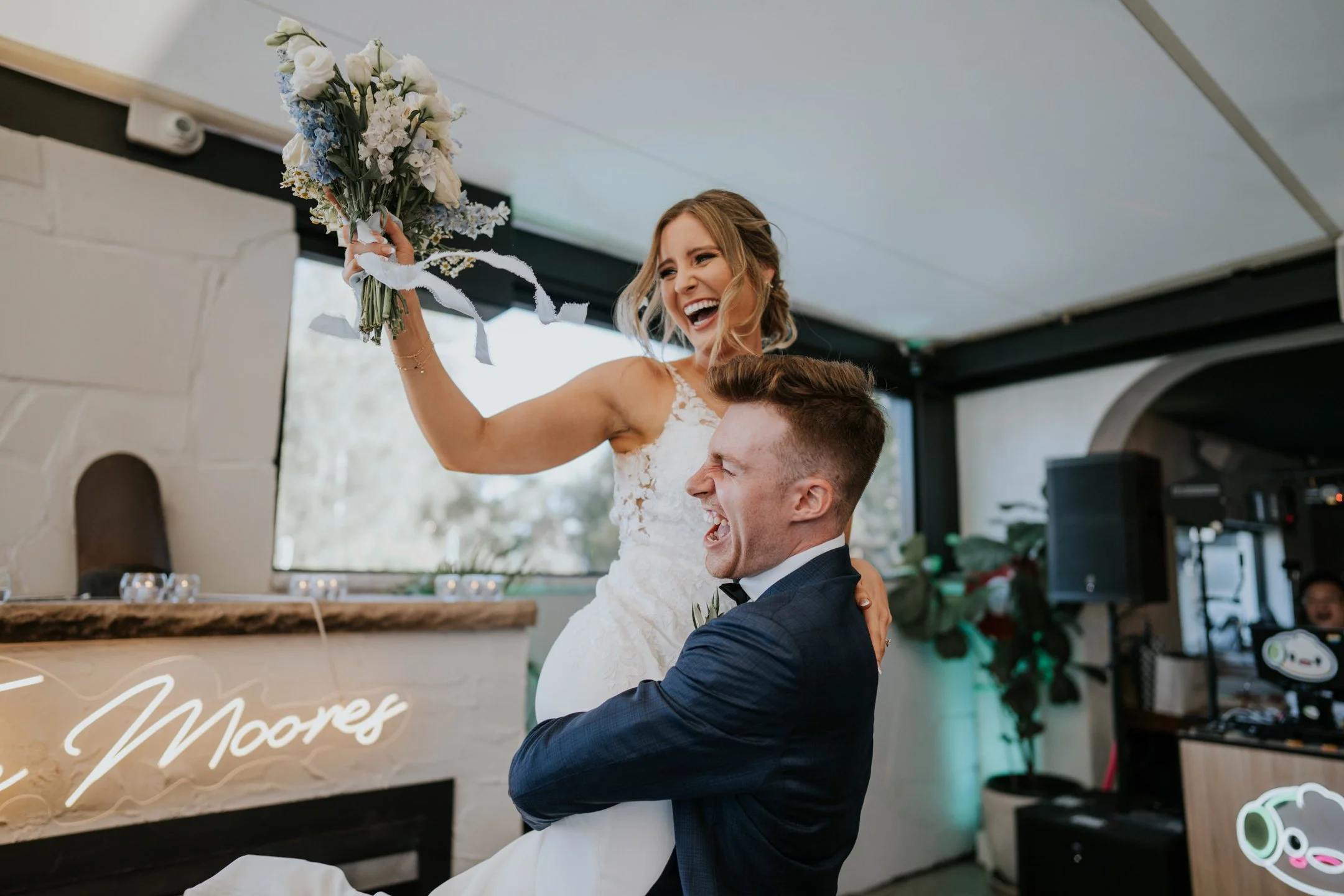 A joyful bride holding a bouquet of flowers is lifted by a groom inside a celebration venue. The couple is smiling and the bride appears excited. The venue has a modern decor with visible lighting and a neon sign.