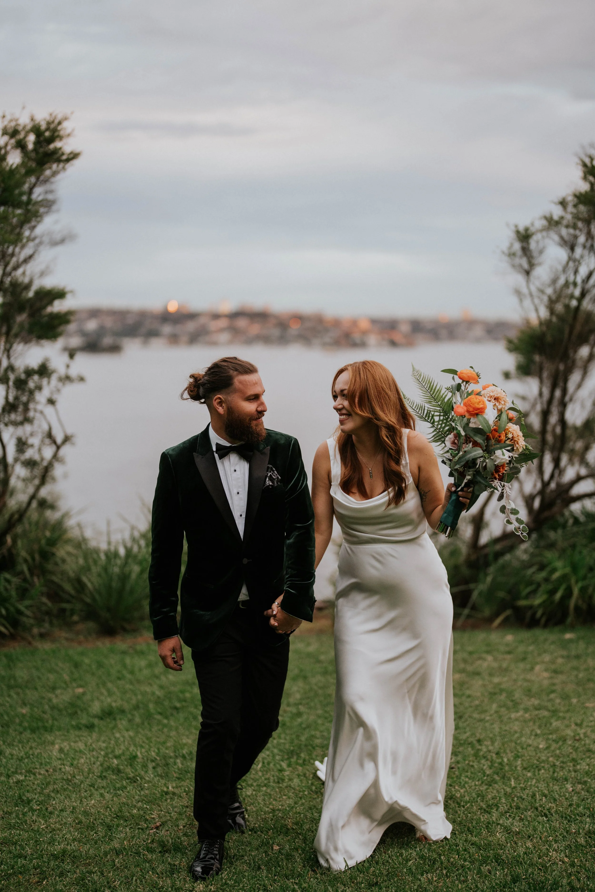 A bride and groom holding hands and walking outdoors near a body of water, smiling, with trees and a cloudy sky in the background. The groom has a beard and is wearing a tuxedo, the bride has red hair and is holding a bouquet of flowers.