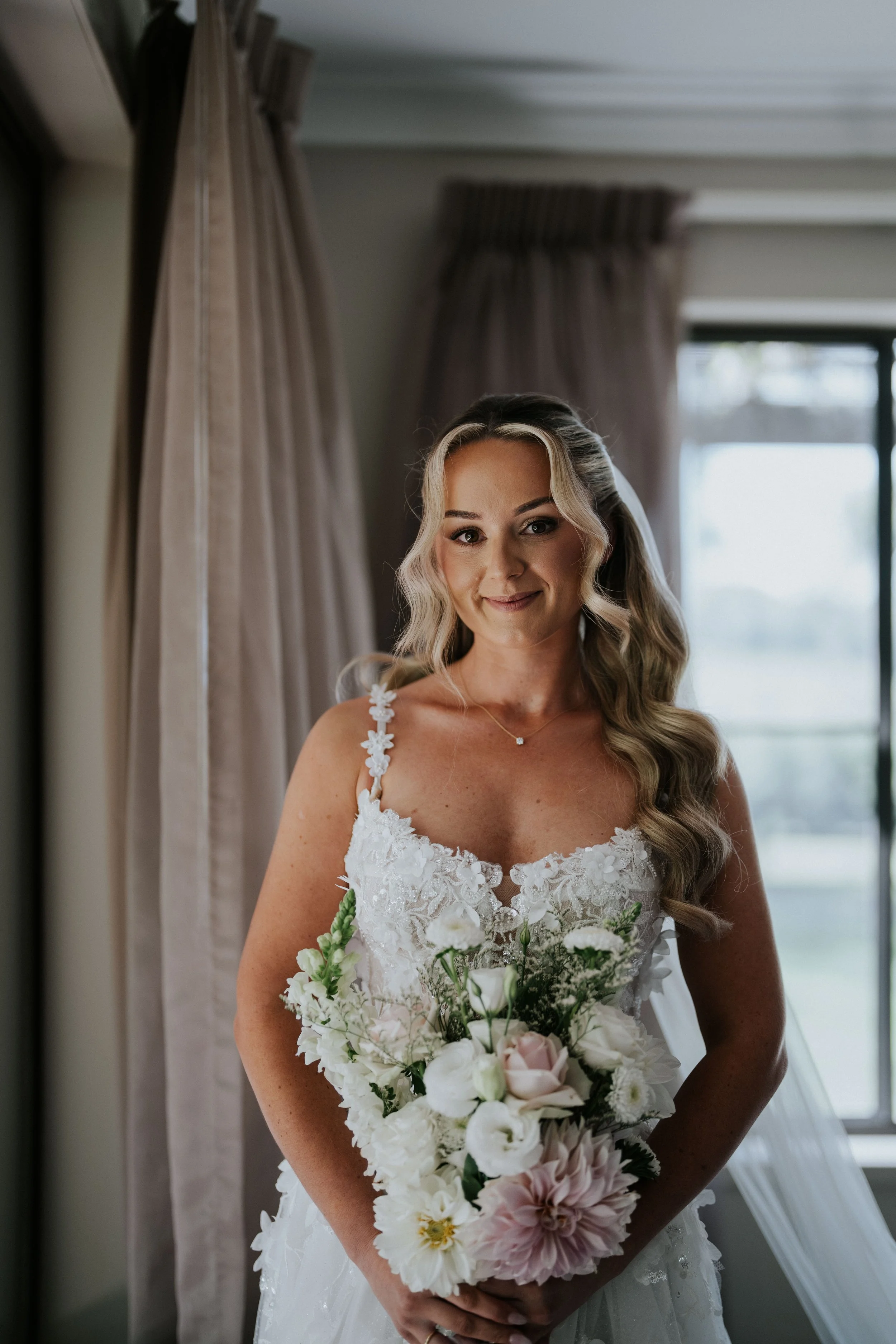 A bride with long wavy hair in a white lace wedding dress, holding a bouquet of white and pink flowers, standing indoors near a window with curtains.