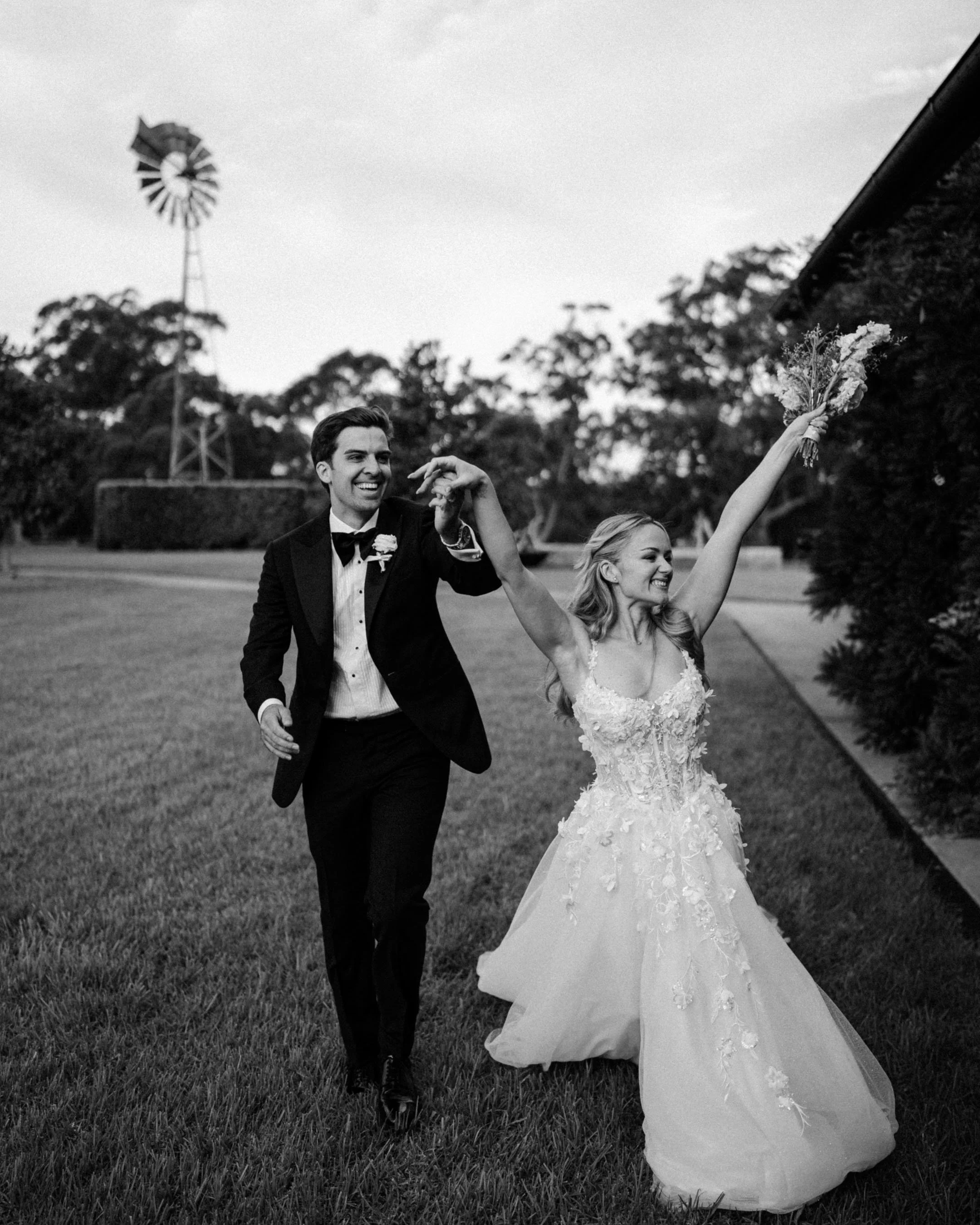 Black and white photo of a wedding couple outdoors. The groom in a tuxedo is smiling and holding the bride's hand. The bride is wearing a lace wedding gown, holding a bouquet, and her other hand is raised.