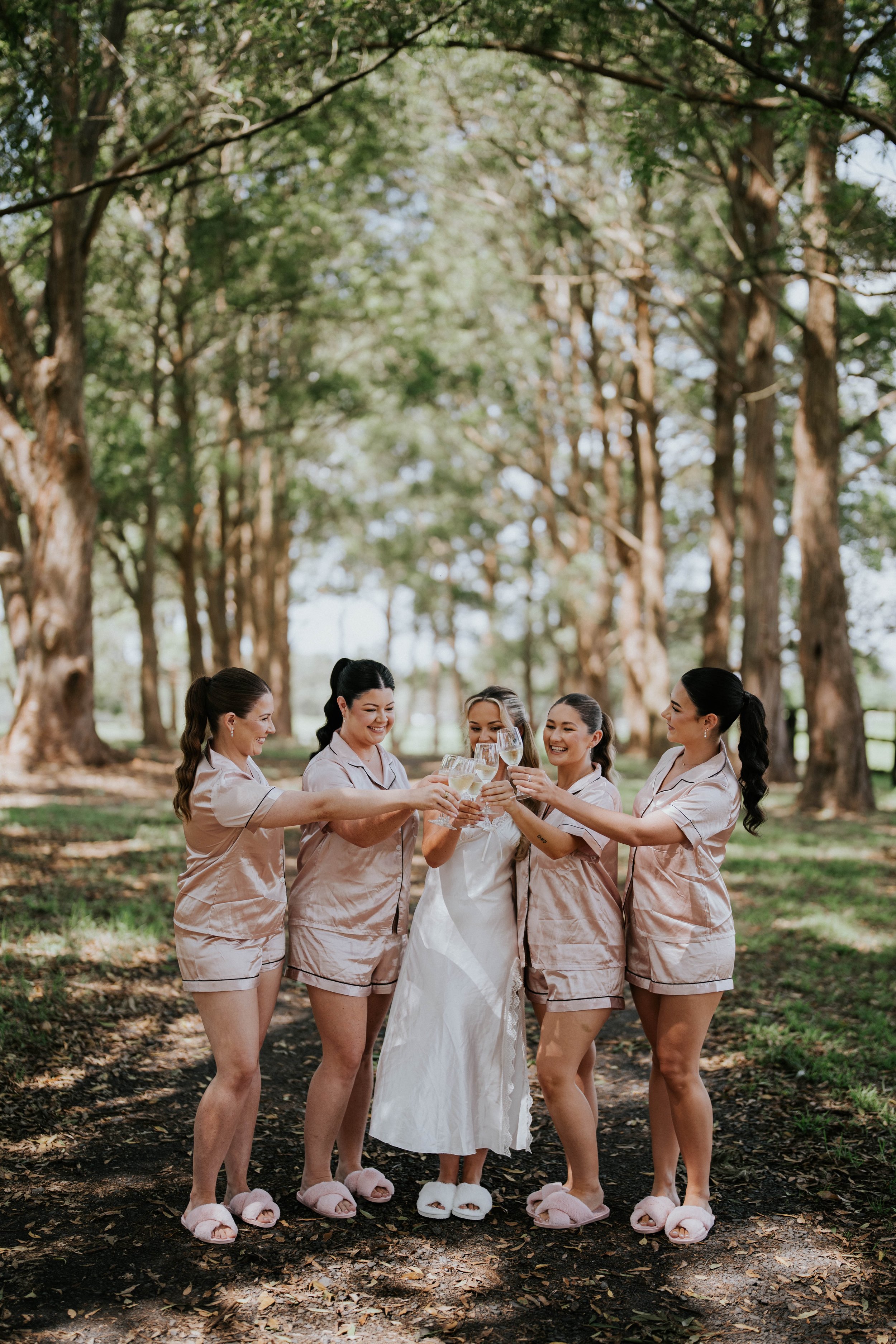 Group of women in pajamas and slippers celebrating with wine glasses in an outdoor park with tall trees.