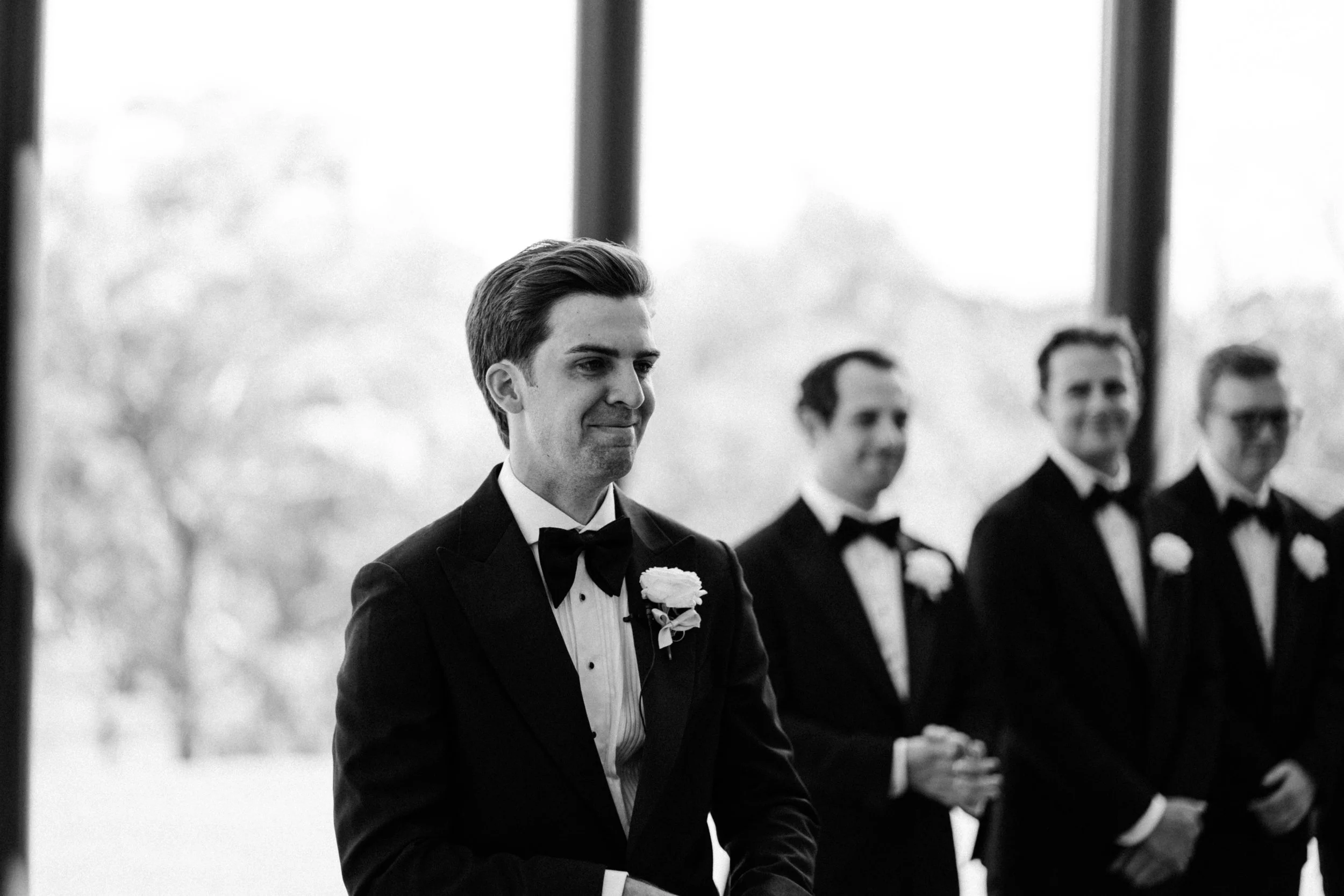 A group of men dressed in tuxedos with bow ties, standing in a line, outdoors during the daytime. The focus is on a man in the foreground with dark hair, smiling subtly, with three other men in the background, all smiling. The image is black and whit