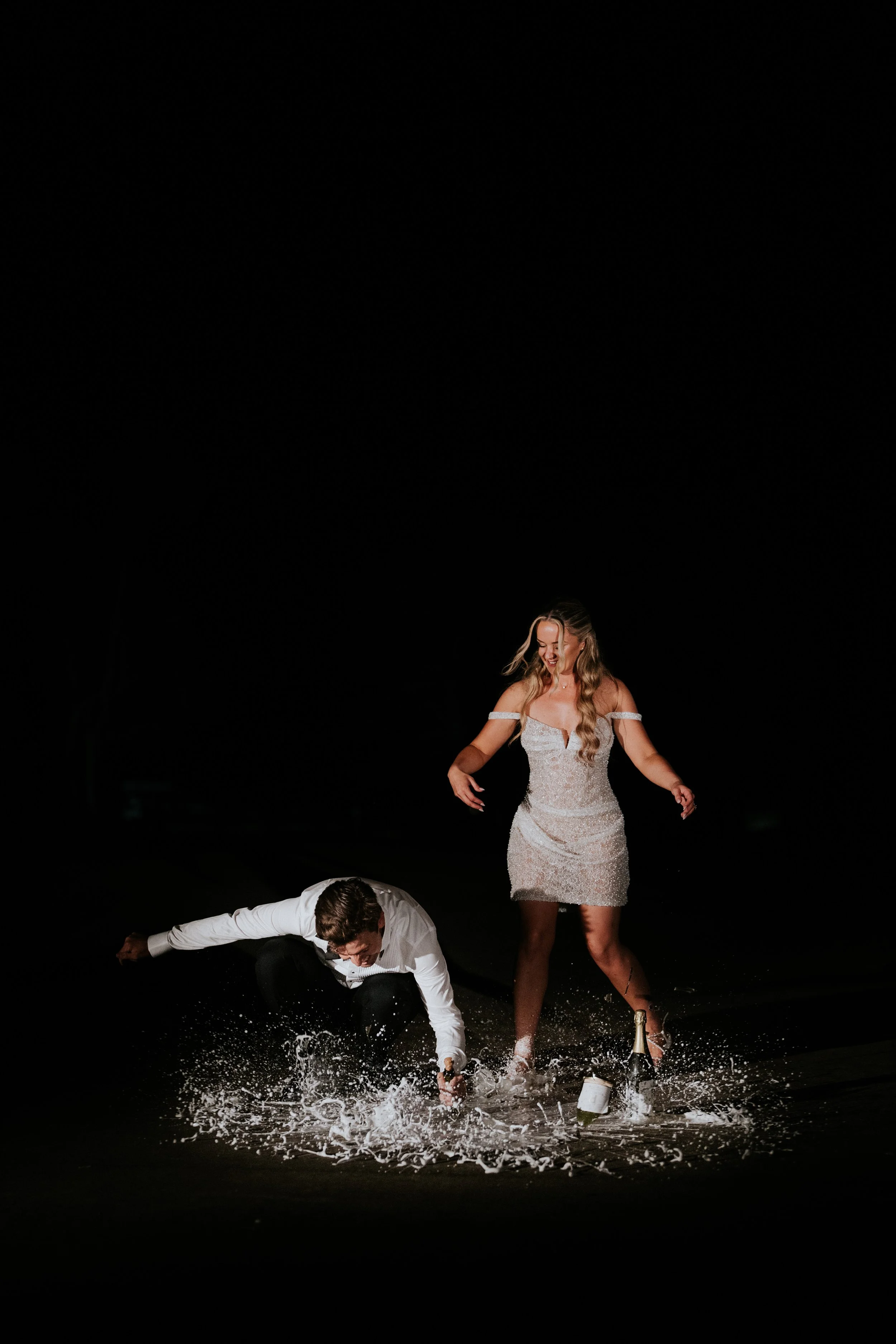 A woman in a white dress is standing on a dark stage, smiling and looking down. A man in a white shirt is falling or has fallen on the stage, with water splashing around him. There is a bottle and other objects on the floor.