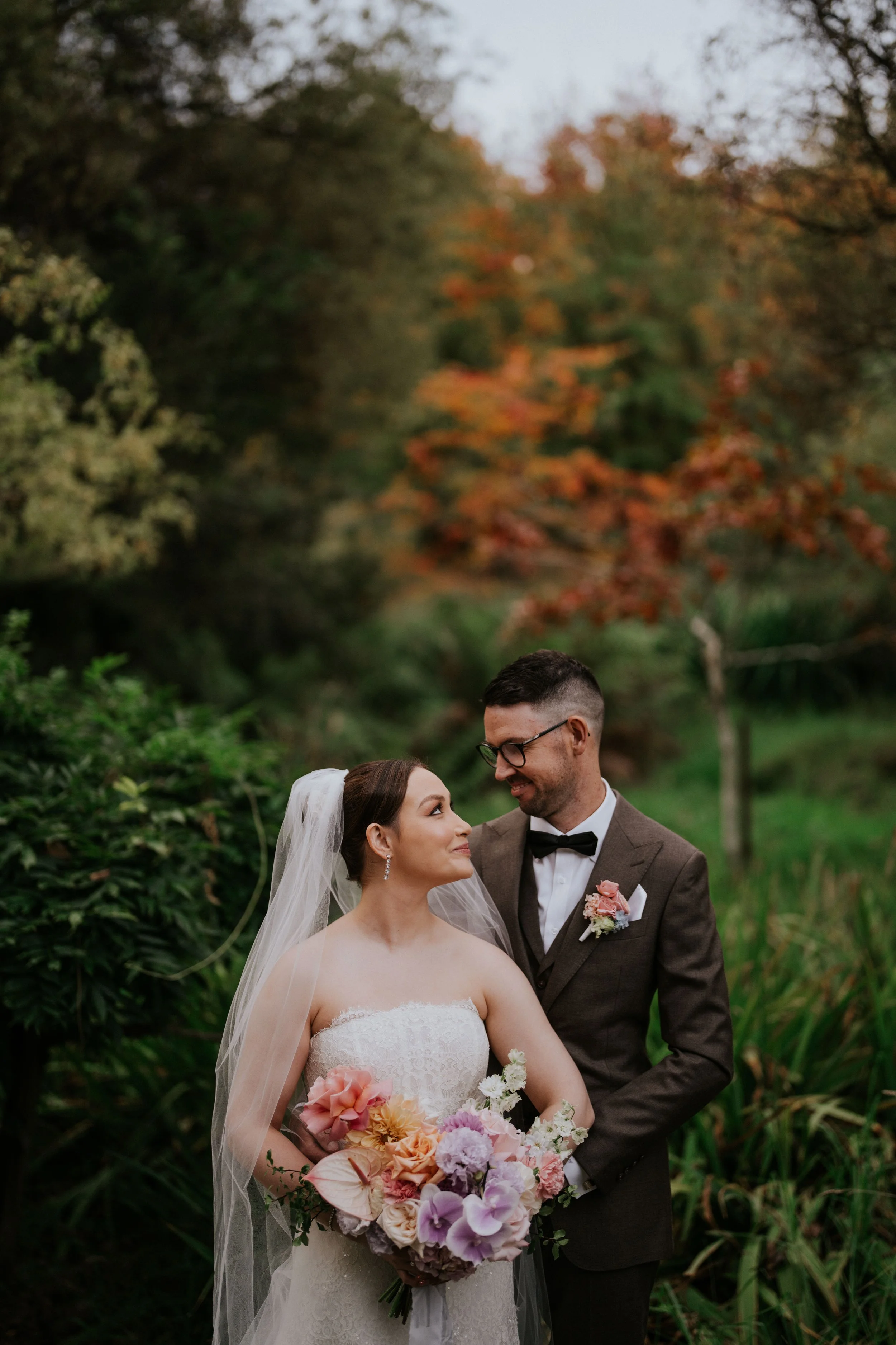 A bride and groom standing close together outdoors with trees and fall foliage in the background. The bride holds a large bouquet of colorful flowers, and the groom is smiling at her while dressed in a brown suit with a bow tie.